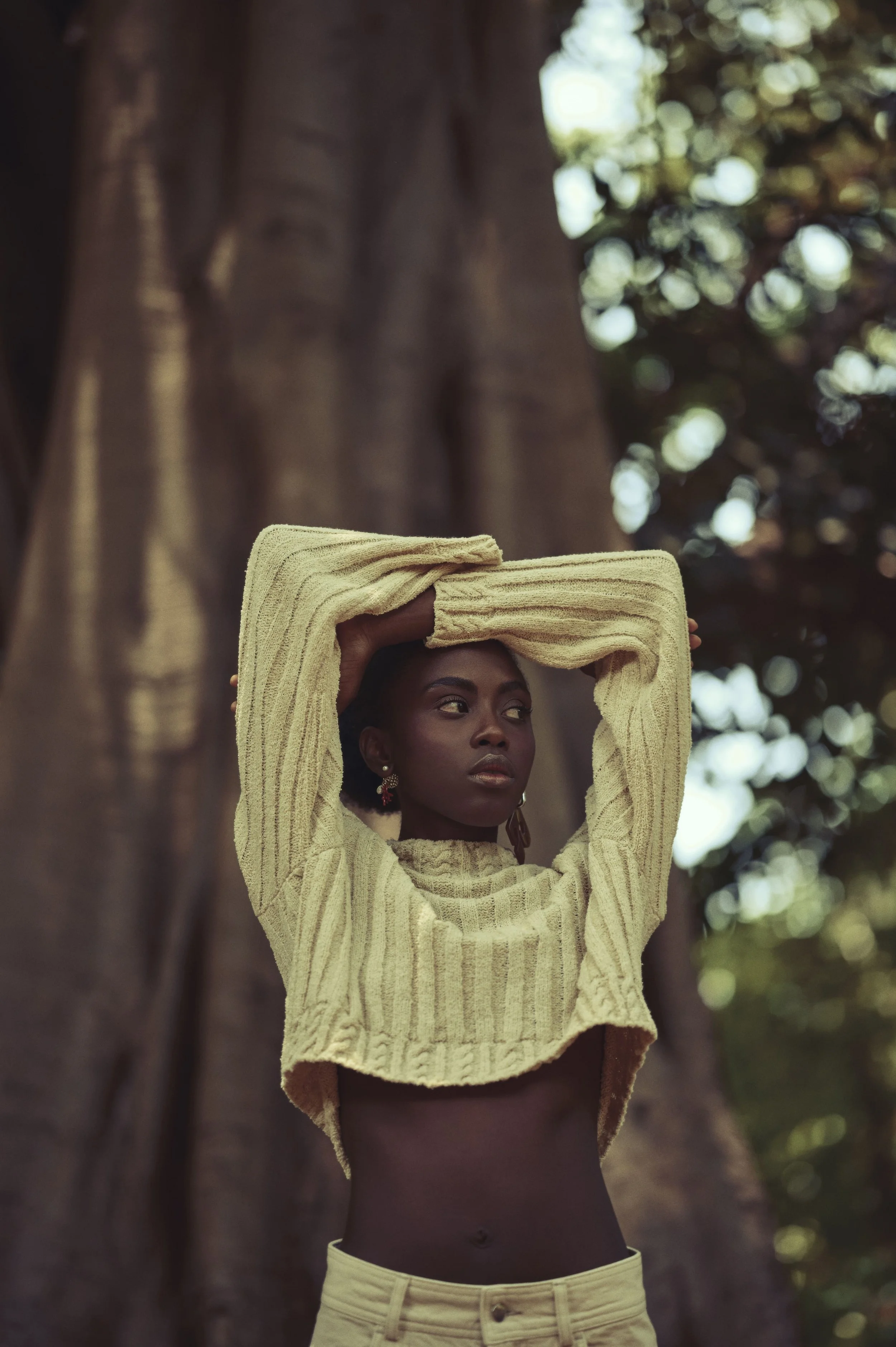 Een vrouw met donkere huid poseert voor een grote boom, ze draagt een gele gebreide trui met lange mouwen en lichtgekleurde broek, met haar armen over haar hoofd, in een natuurlijke omgeving in Cuba gefotografeerd door Eric van den Elsen.