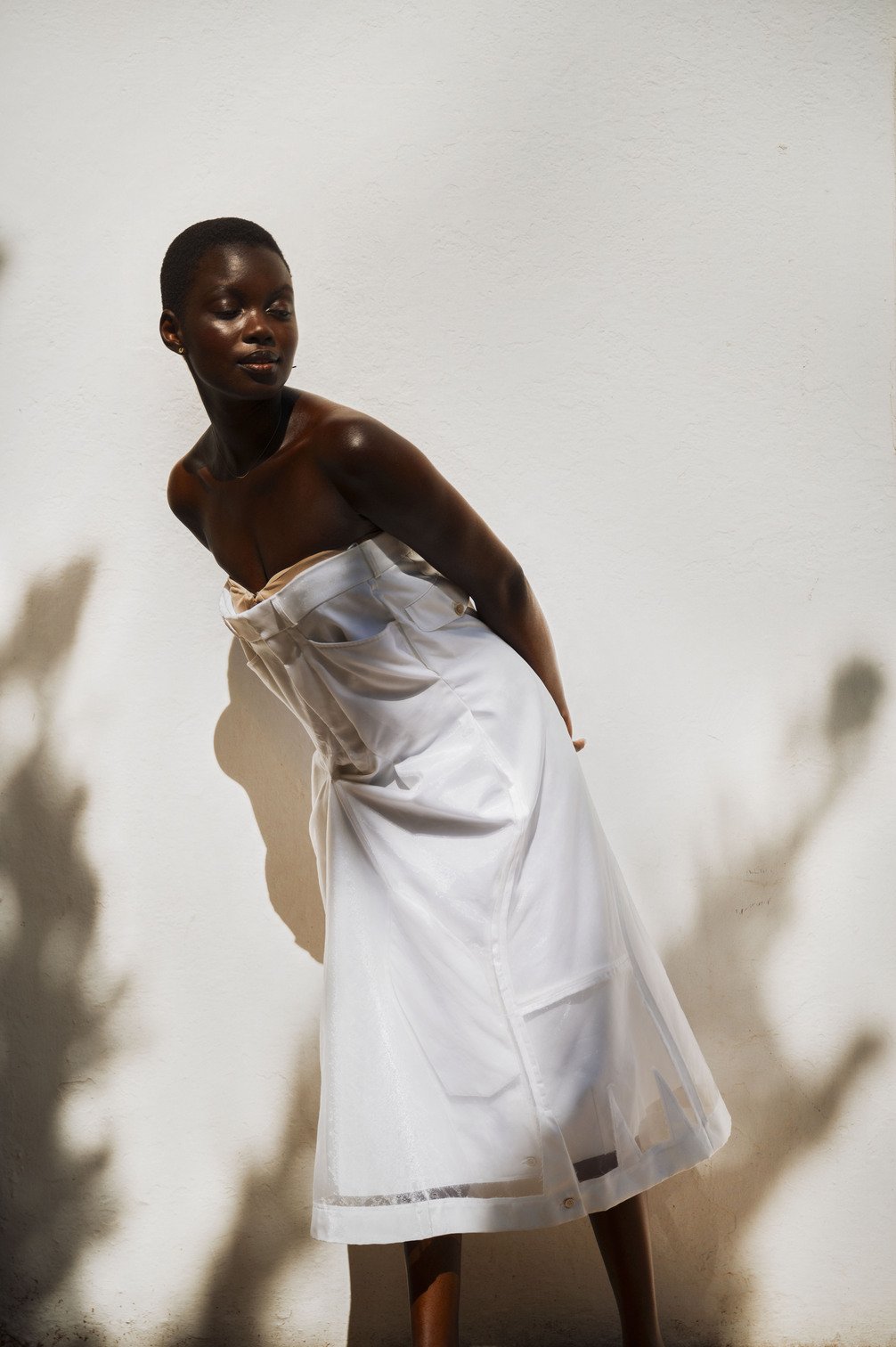 Een vrouw met een gebruinde huid die op een witte muur staat, gekleed in een wit satijnen rokje gefotografeerd door Eric van den Elsen.