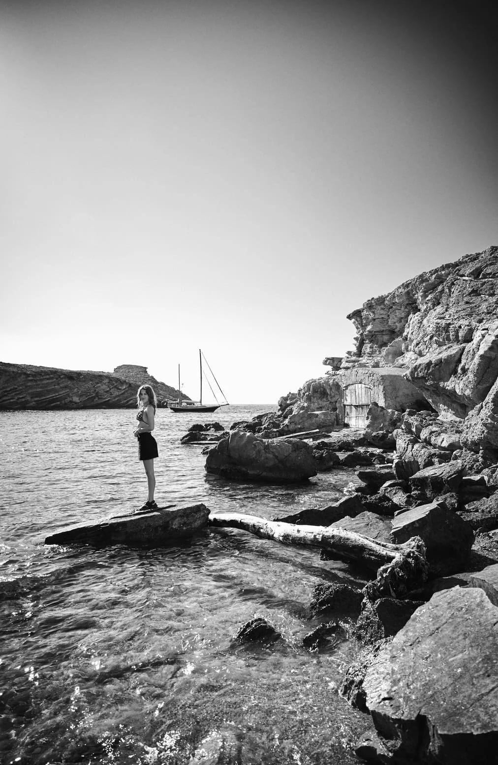 Een zwart-wit foto van een vrouw die op een rots in de zee staat, met een nabijgelegen rotswand en een zeilboot op het water. De achtergrond toont een rieteiland en een heldere, lege lucht.