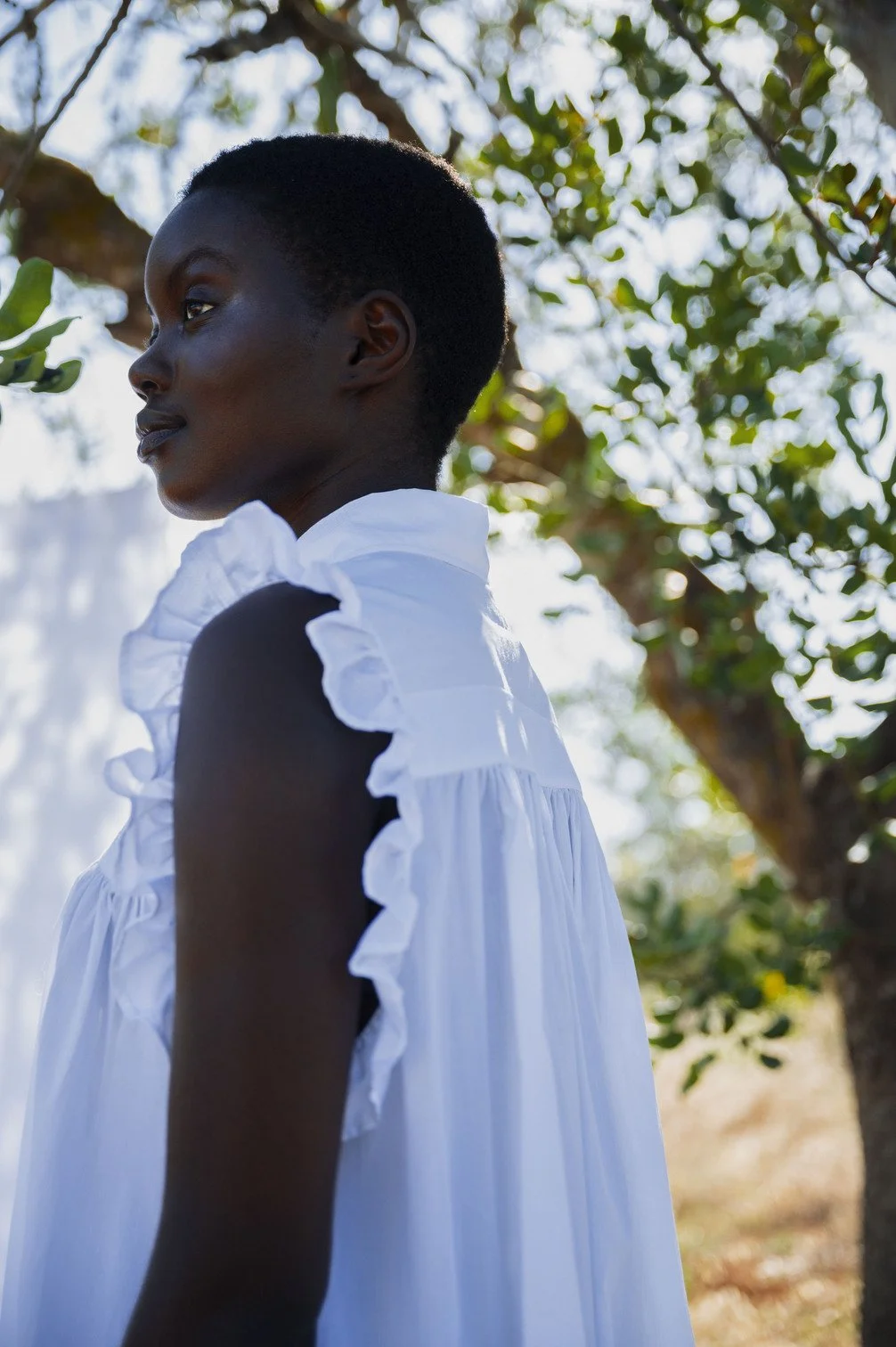 Portret van een vrouw met kort zwart haar in een witte jurk buiten, omgeven door groene bladeren en bomen gefotografeerd door Eric van den Elsen.