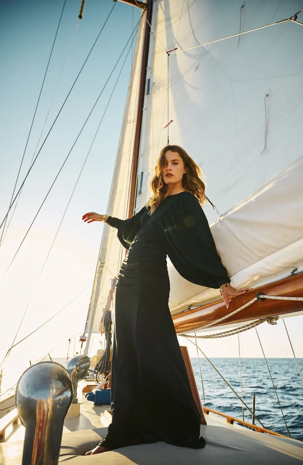 Vrouw in een lange zwarte jurk poseren op een jacht, met de zee en de horizon op de achtergrond, tijdens zonsondergang.