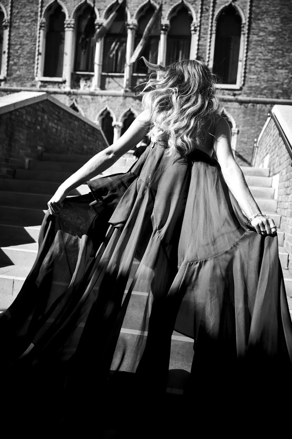 Een vrouw met golvend haar in een lange jurk die op trappen staat, met een historische gebouw op de achtergrond, in zwart-wit gefotografeerd door Eric van den Elsen.