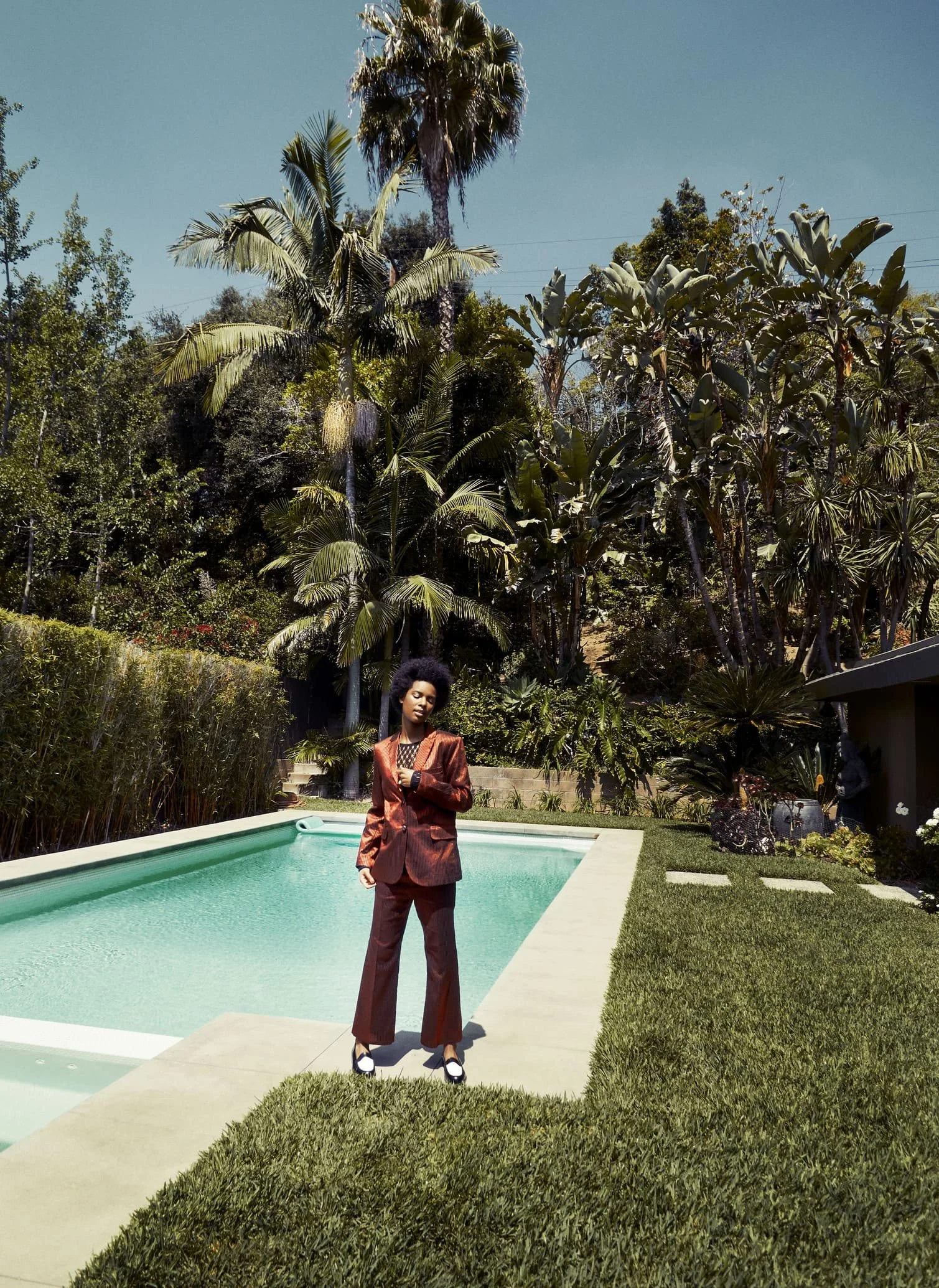 Een vrouw in een bruin pak staat naast een zwembad, omgeven door palmbomen en tropische planten op een zonnige dag gefotografeerd door Eric van den Elsen.