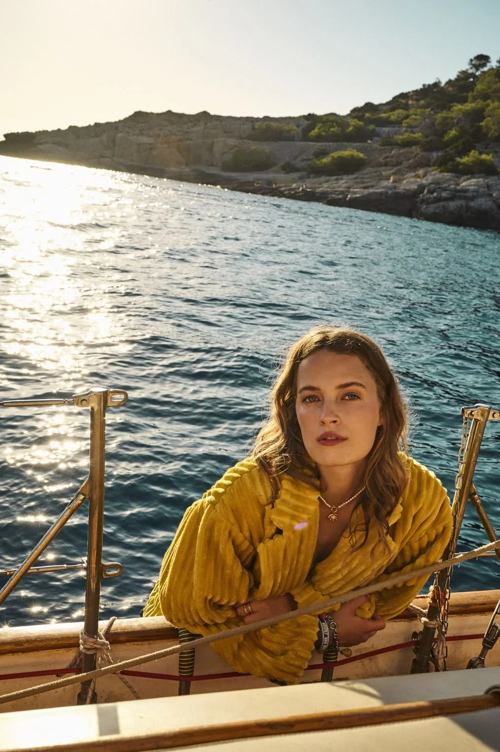 Een jonge vrouw met bruin haar zittend op een boot met blauwe wateren eromheen en een rotsachtig landschap op de achtergrond, tijdens zonsondergang.
