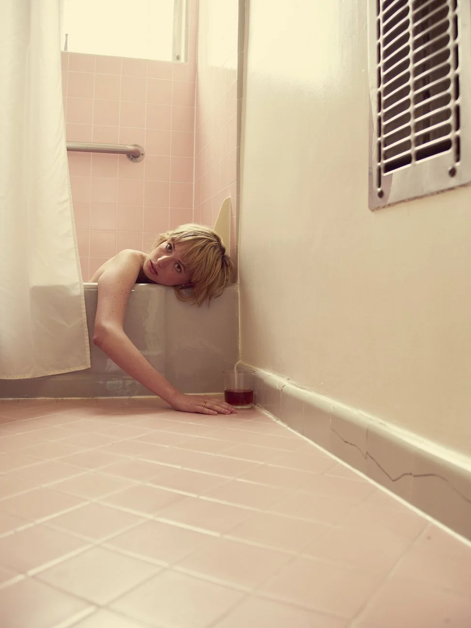 Persoon in een badkuip met het hoofd buiten de douche, die over de rand leunt en naar de camera kijkt, met een glas cola op de vloer gefotografeerd door Eric van den Elsen.