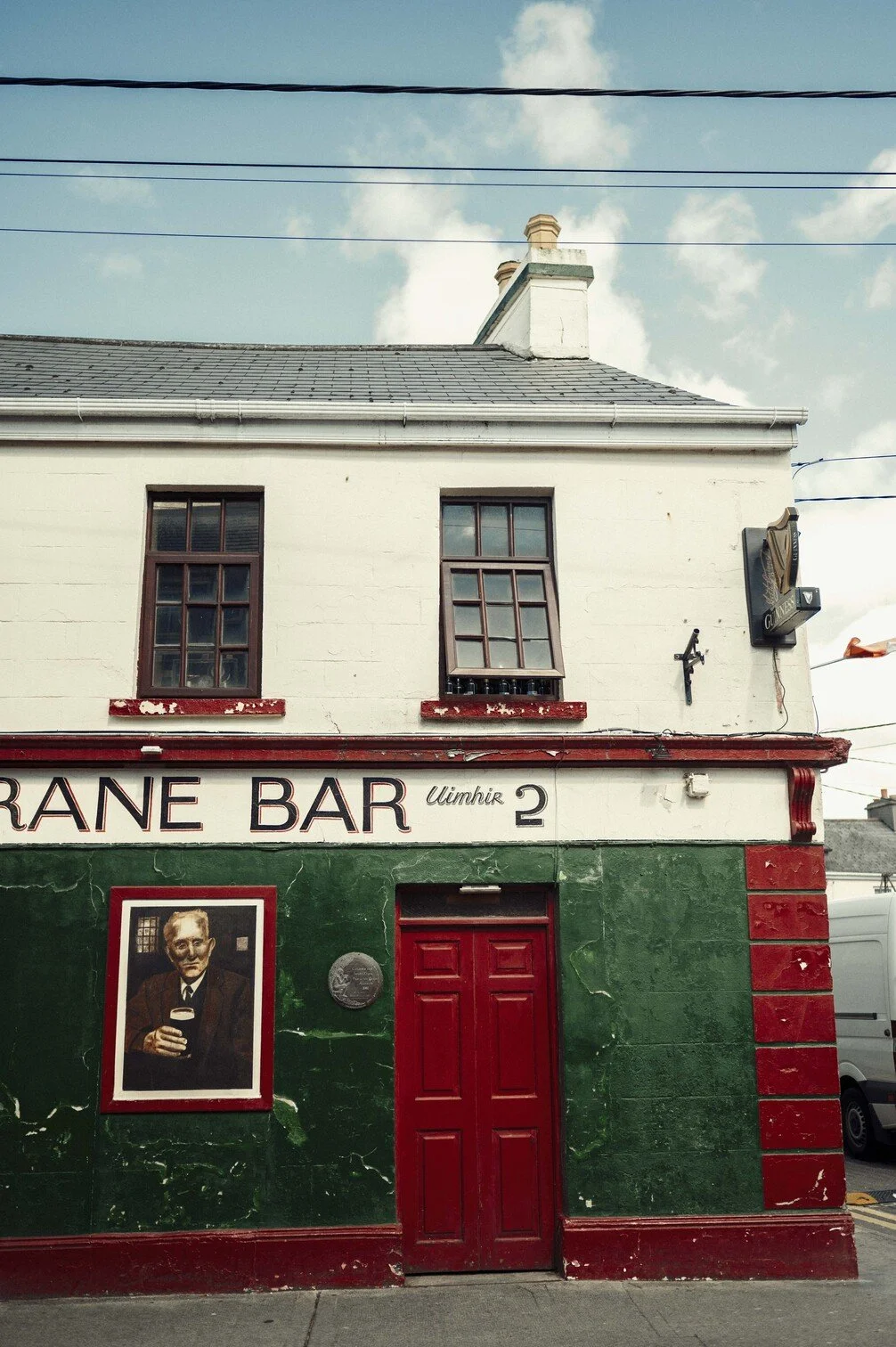 Een café of bar met een witte bovenkant en een groene onderkant, met een rood deur en raamkozijnen. Een schilderij van een man met een glas in de hand hangt aan de muur gefotografeerd door Eric van den Elsen.