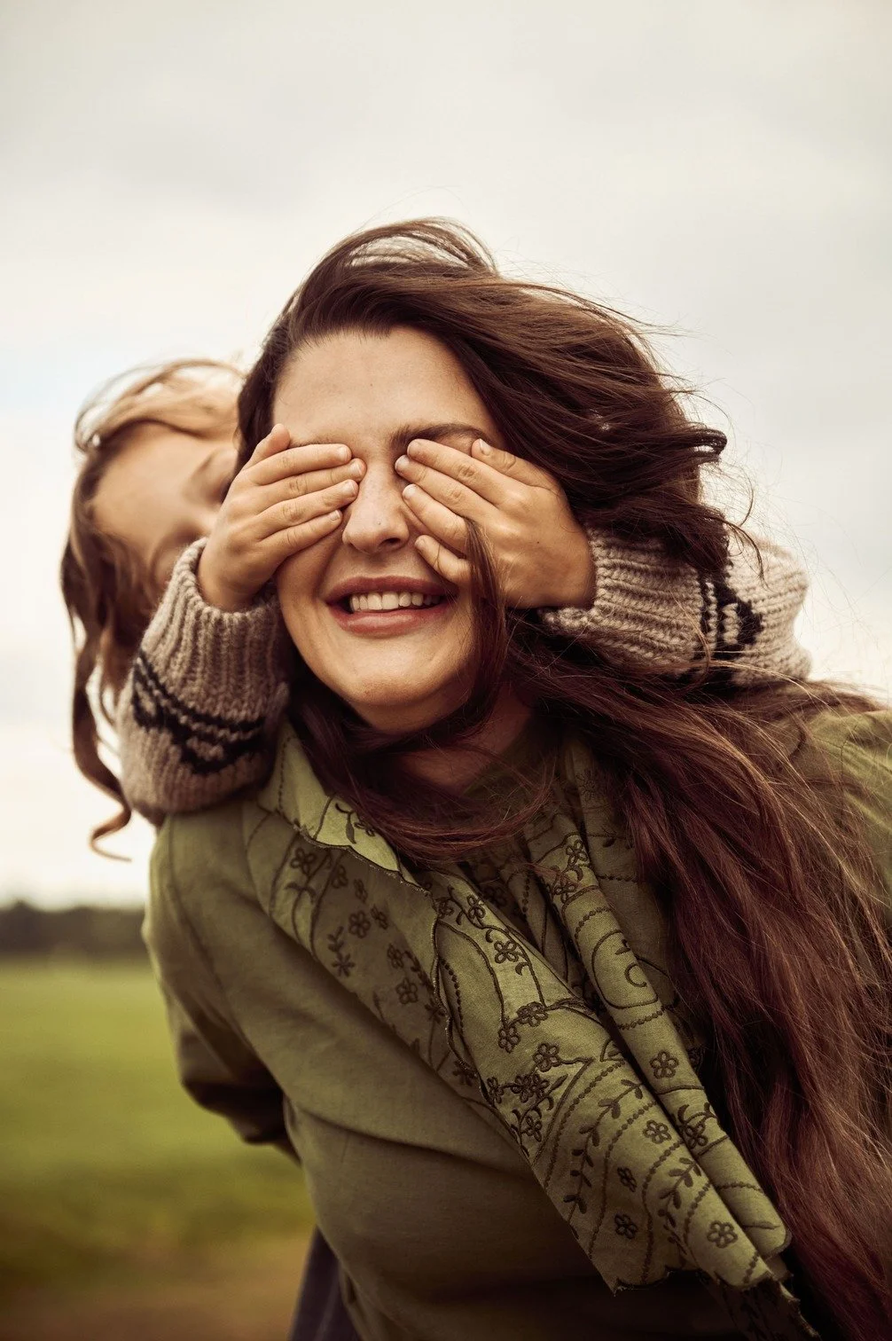 Romy Boomsma met lang bruin haar krijgt handen over haar hoofd door haar meisje die haar ogen bedekt met haar handen in een veld met een bewolkte lucht gefotografeerd door Eric van den Elsen.