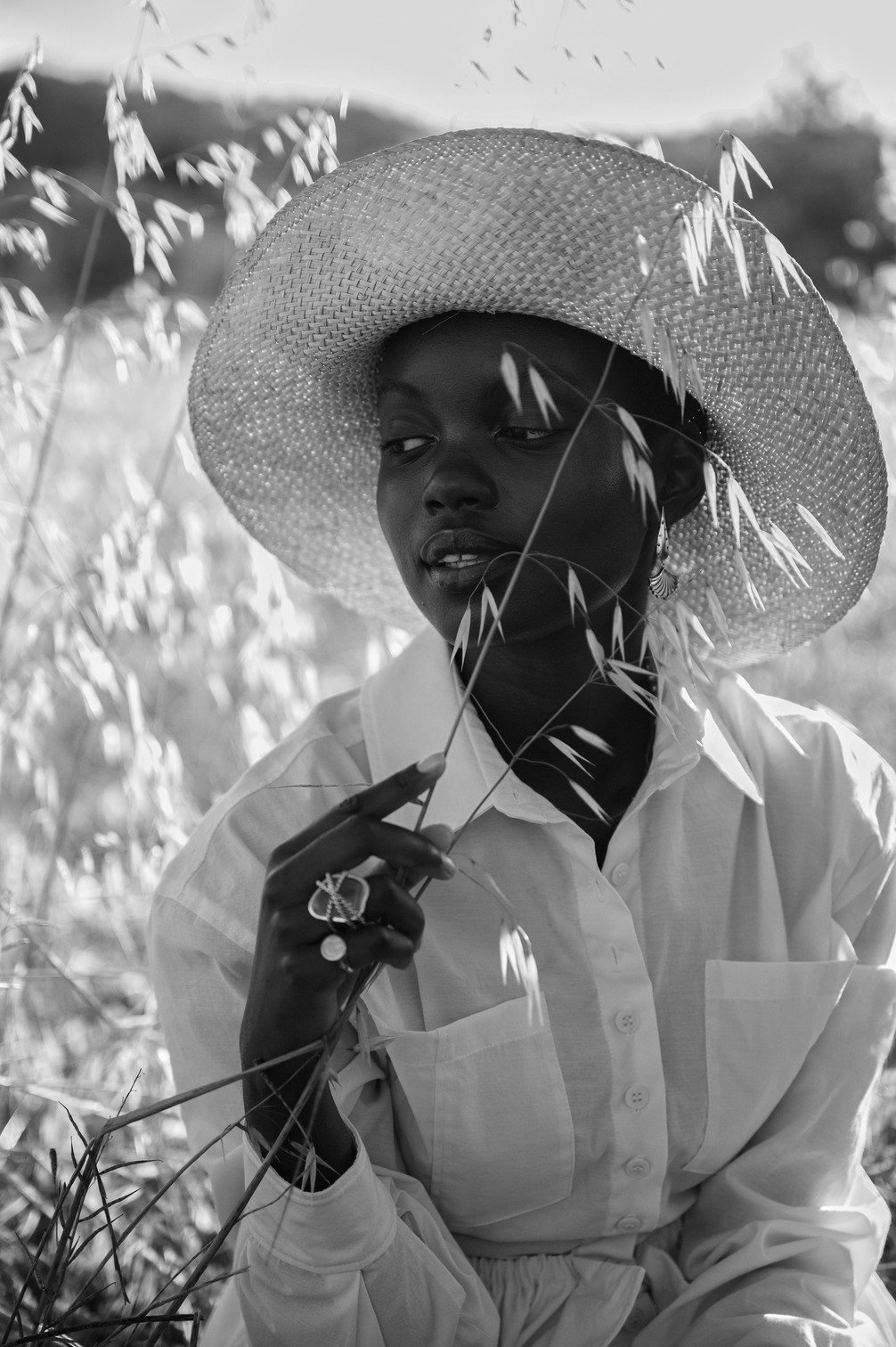 Zwarte vrouw met een grote strandrook, witte blouse en sieraden, die door gras of gewassen kijkt gefotografeerd door Eric van den Elsen.