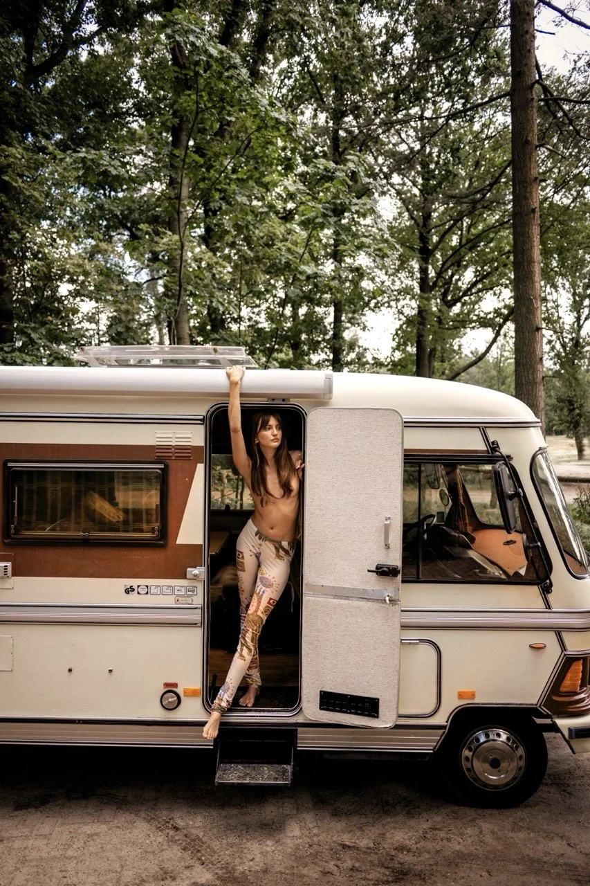 Vrouw met lang haar en geen shirt, staand in de deuropening van een oude kampeerauto, met één arm omhoog en de andere vasthoudend aan de dakrand, in een bosrijke omgeving gefotografeerd door Eric van den Elsen.