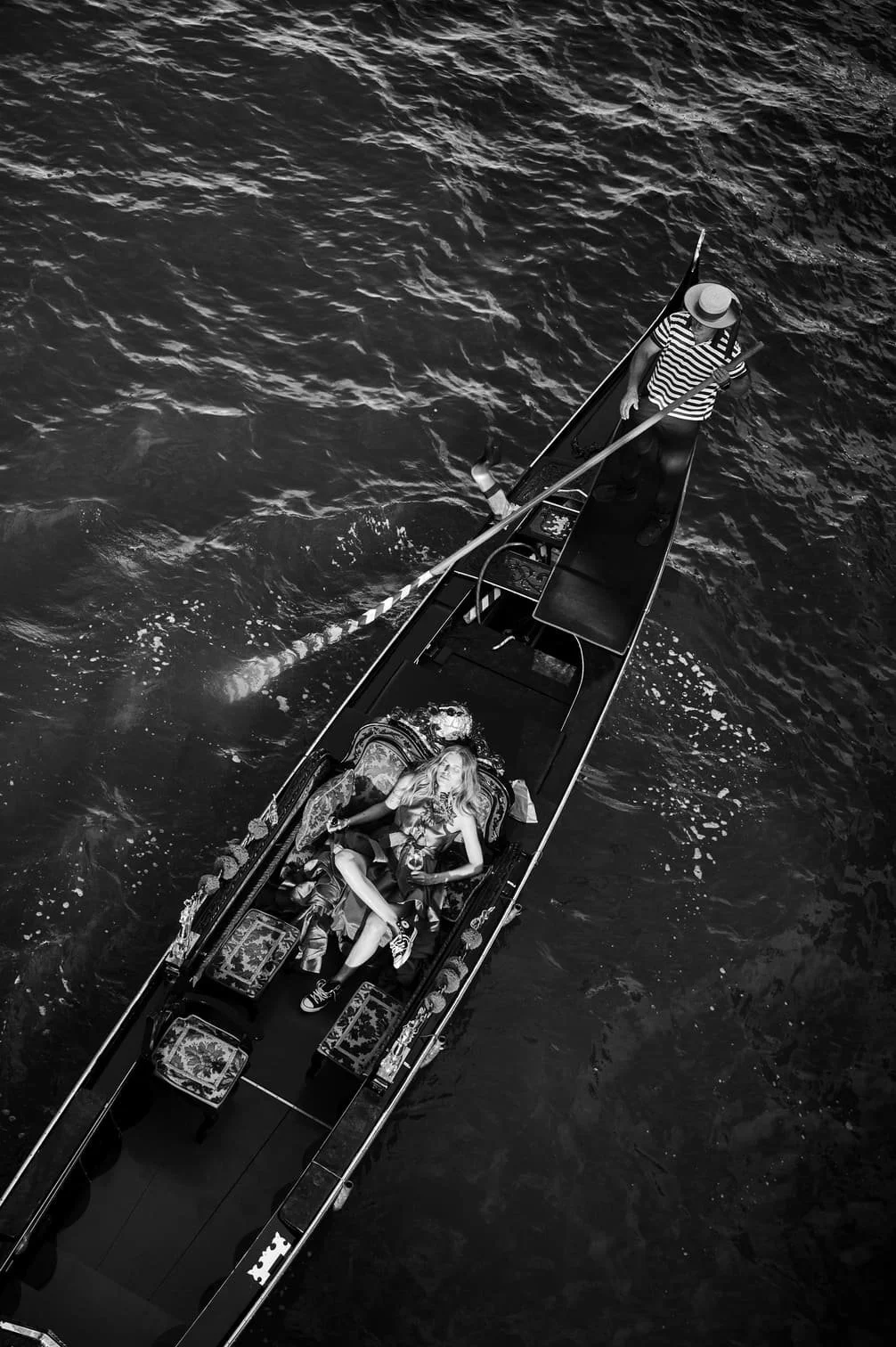 Een zwart-wit foto van een draaimolenboot op het water met een vrouw die op een driekwartstoel ligt en een man die roeiriemen vasthoudt, gezien van boven gefotografeerd door Eric van den Elsen.
