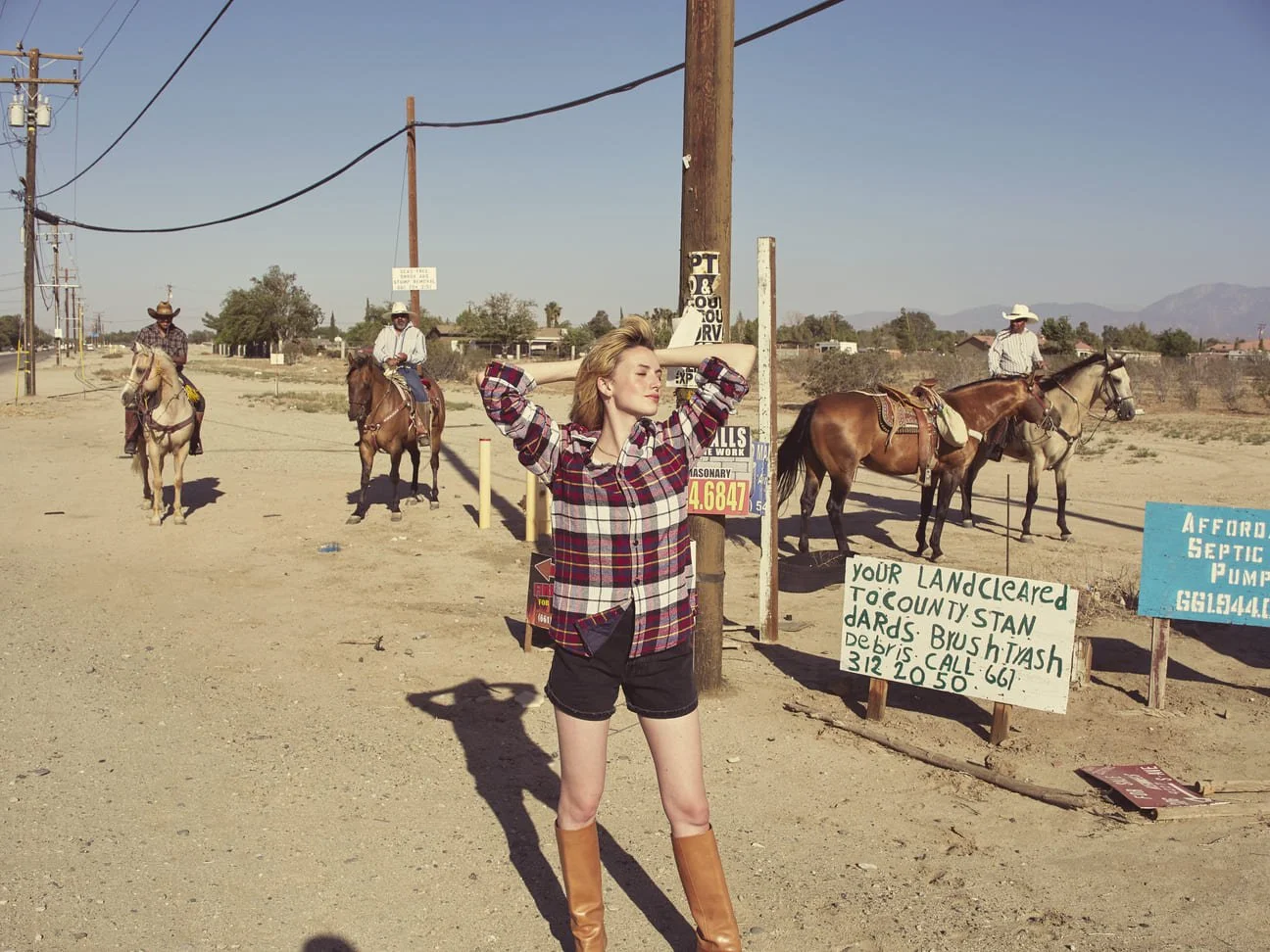 Een meisje poseert met haar ogen dicht en handen achter haar hoofd op een stoffige straat met vier ruiters op paarden op de achtergrond gefotografeerd door Eric van den Elsen.