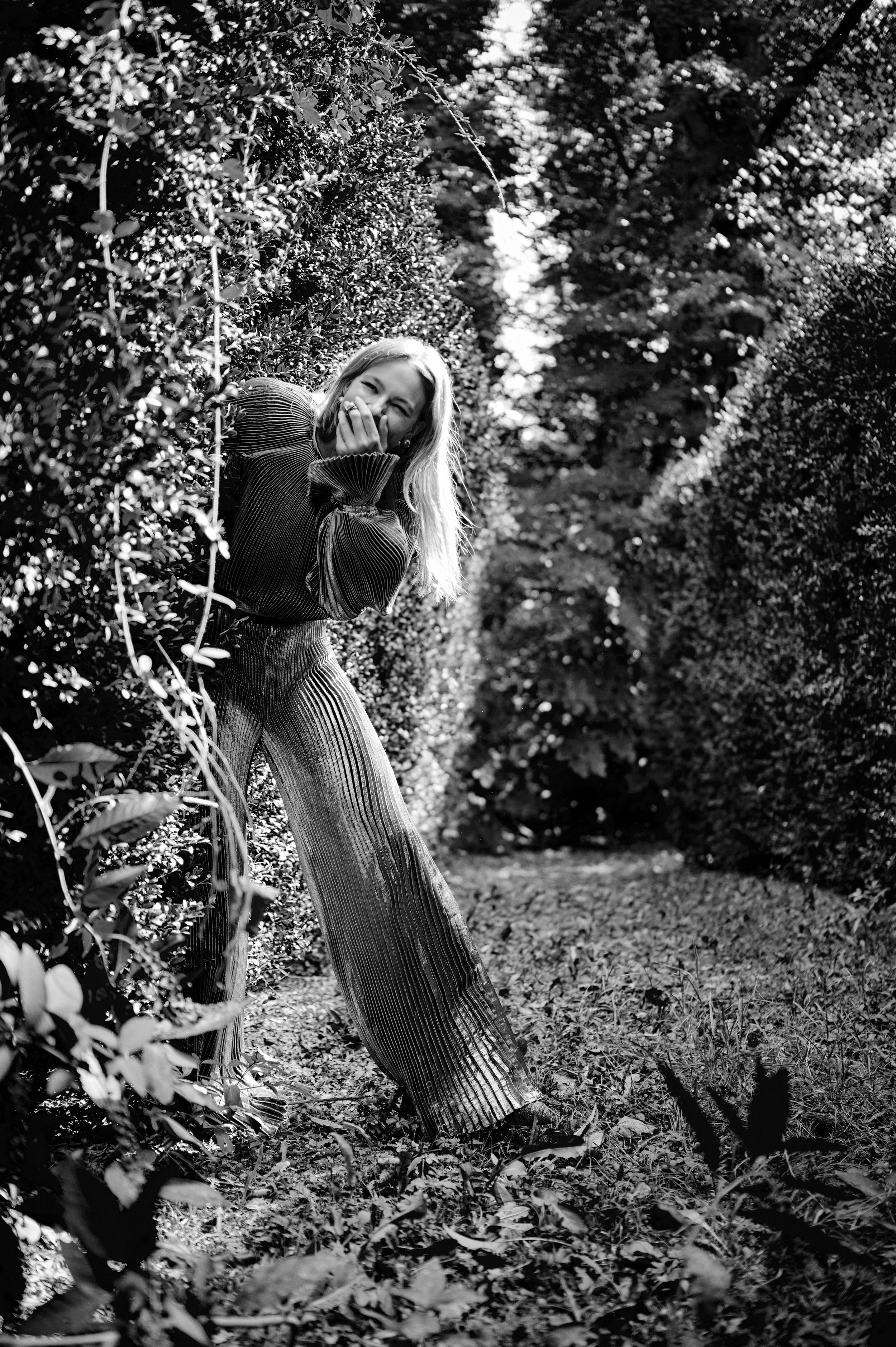 Een lachende vrouw met lang haar in een tuin, omringd door hoog opschietende heggen en bomen, gekleed in een geribbelde, wijde broek en een top, die zich achter een struik verstopt gefotografeerd door Eric van den Elsen.