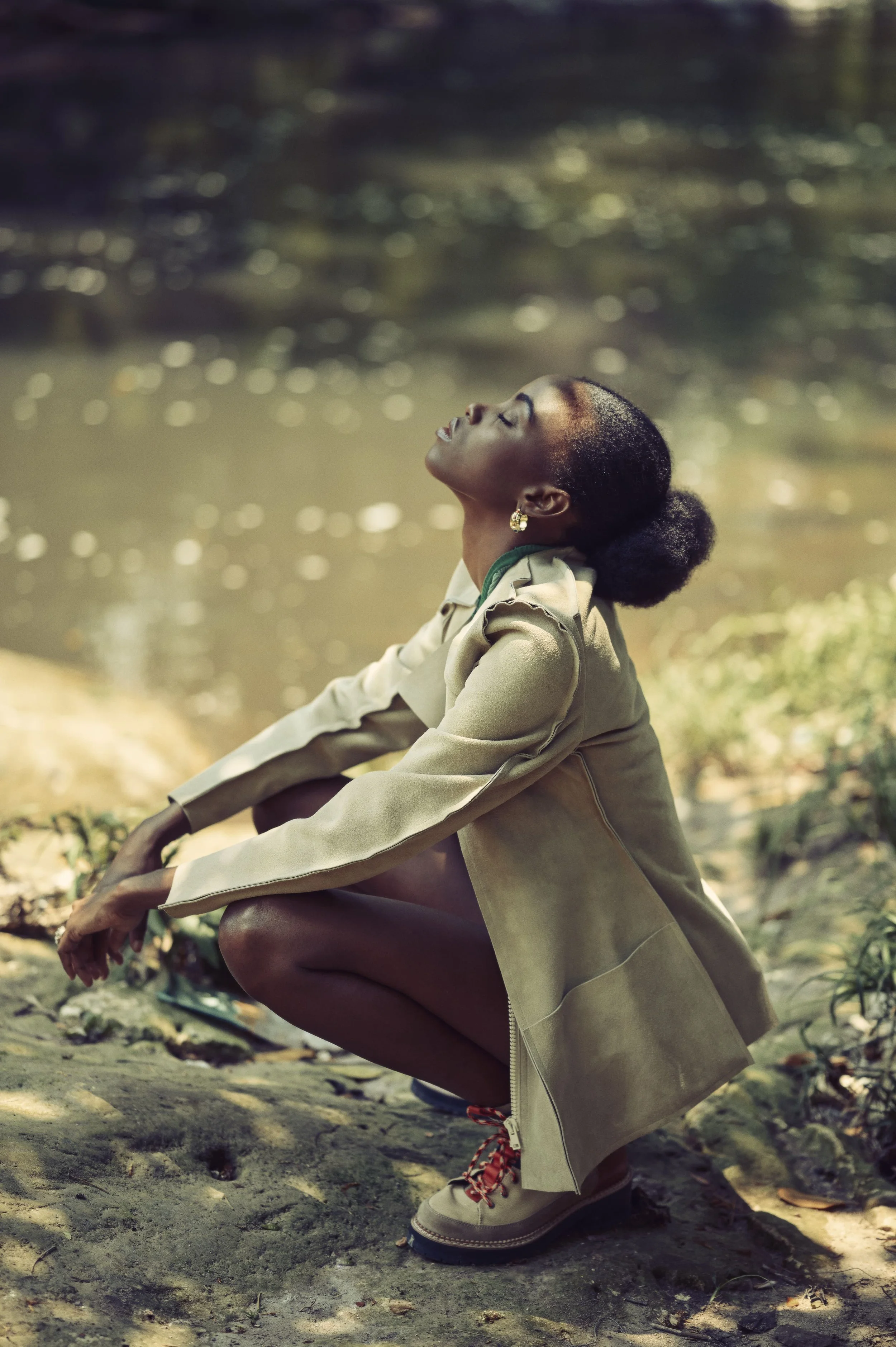 Een vrouw in een beige jas knielt op de oever van een rivier met haar hoofd omhoog en ogen dicht, omringd door een natuurlijke omgeving gefotografeerd door Eric van den Elsen.