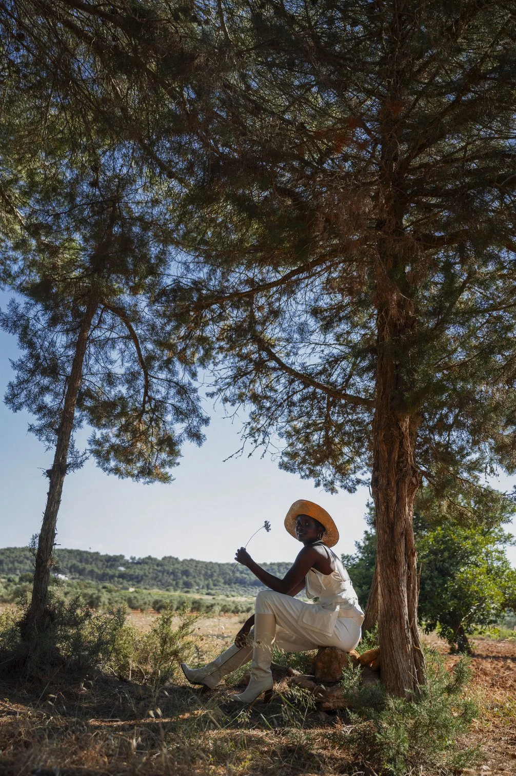 Een vrouw zit onder een grote dennenboom in een veld, draagt witte kleding en een hoed en houdt een kleine bloem vast gefotografeerd door Eric van den Elsen.