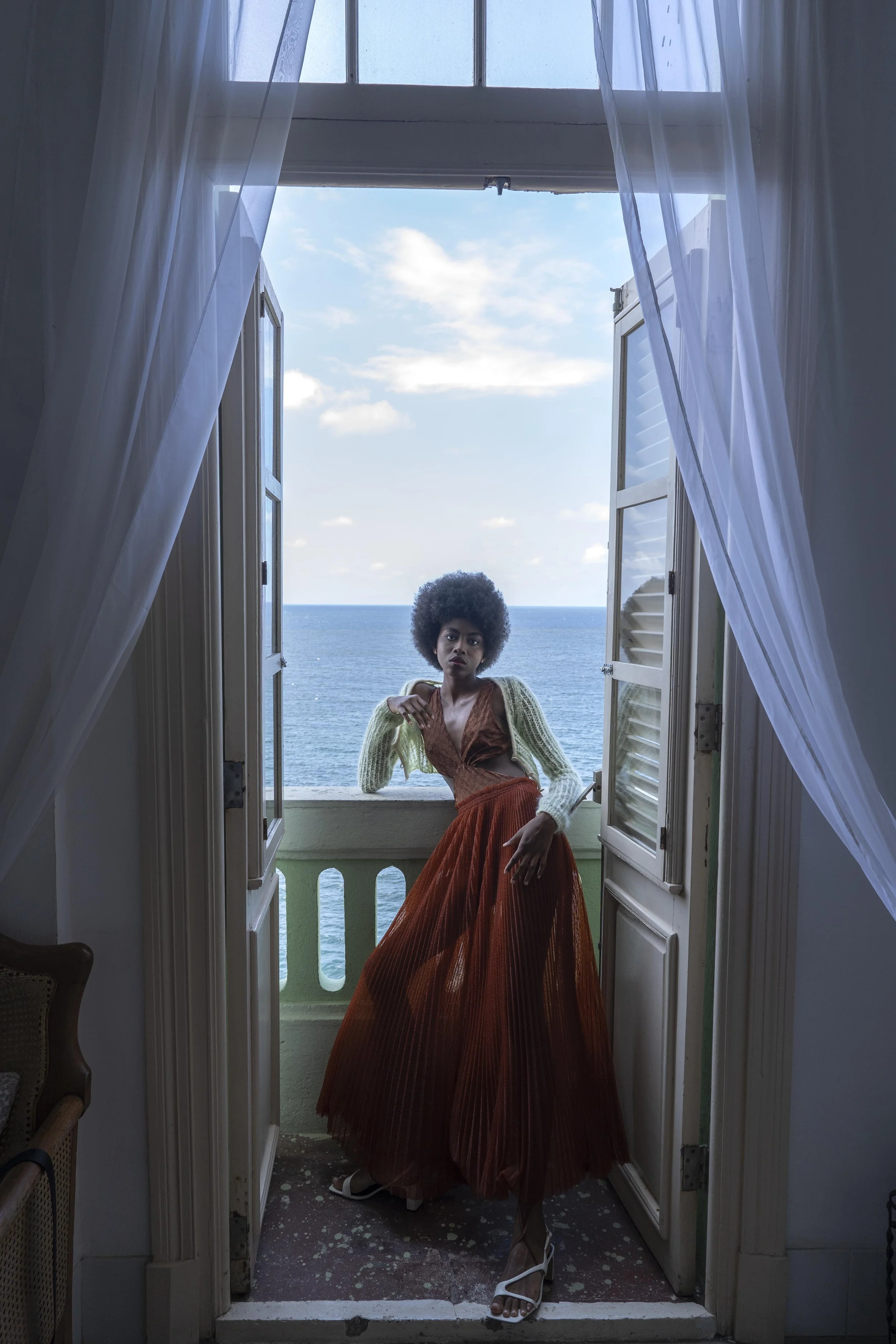 Een vrouw met een grote afro-haar staan op een balkon met uitzicht op de oceaan, binnen in een huis met open ramen en lichte gordijnen gefotografeerd door Eric van den Elsen.