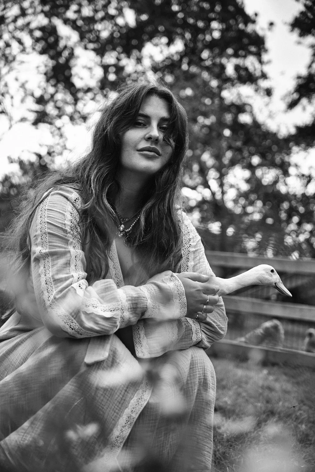 Zwart-wit foto van Romy Boomsma die in een buitenomgeving zit, haar hand rust op een zwanengezichthout. Ze heeft lang, golvend haar en draagt sieraden gefotografeerd door Eric van den Elsen.