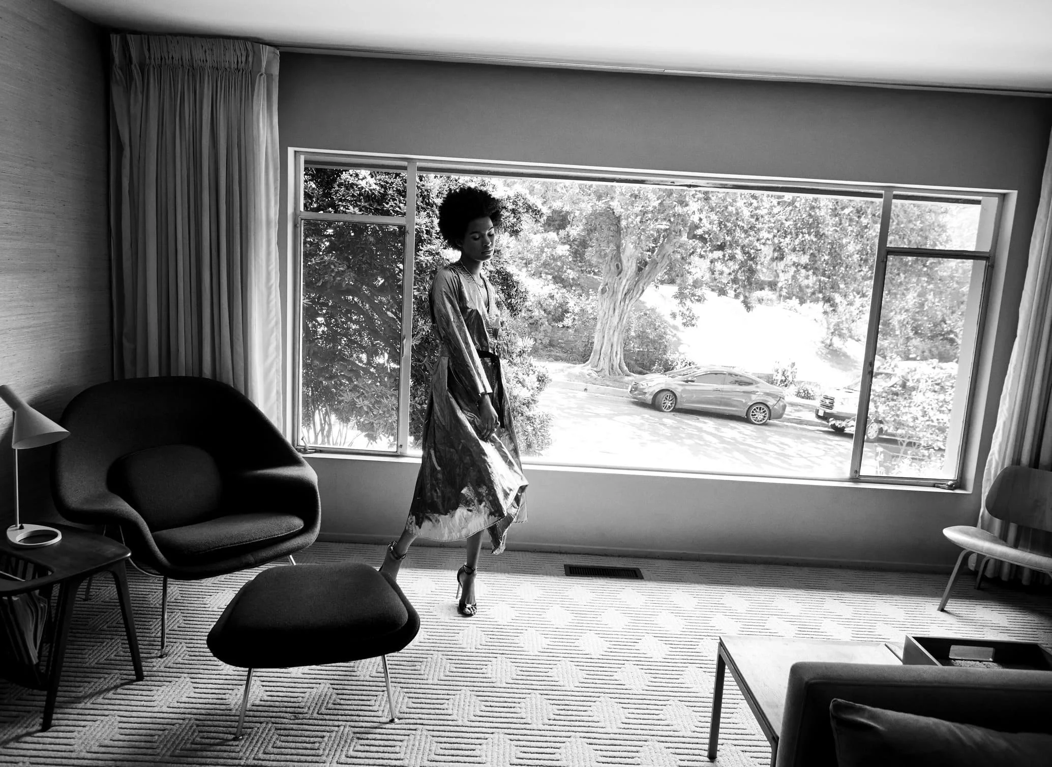 Een zwart-wit foto van een vrouw die in een hotelkamer staat, voor een groot raam met een tuin en geparkeerde auto's zichtbaar buiten gefotografeerd door Eric van den Elsen.