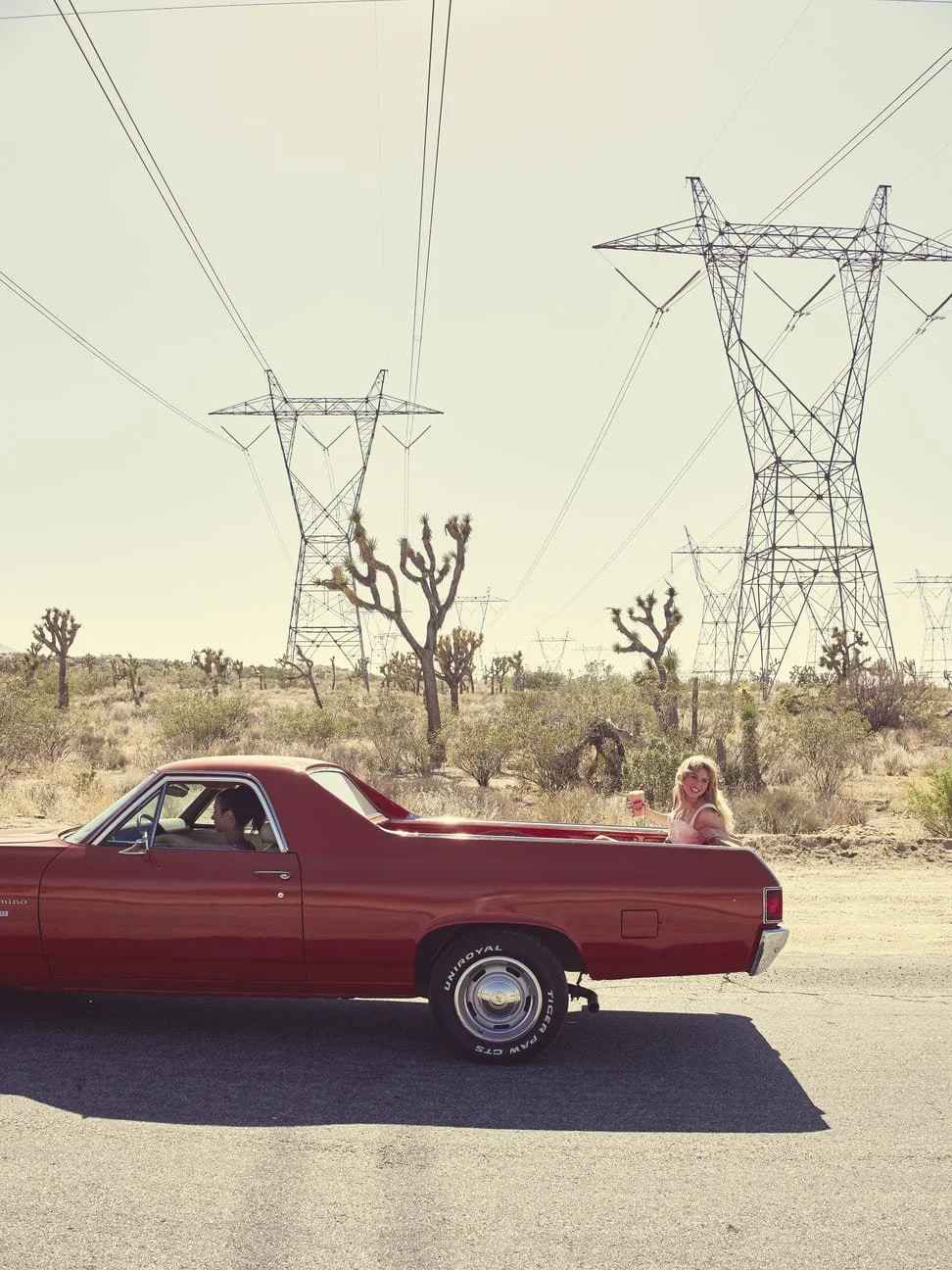 Een rode klassieke pick-up truck rijdt door een woestijnlandschap met cactussen en hoge elektrische hoogspanningsmasten. In de laadbak zit een vrouw die lachend een drankje vasthoudt, terwijl de bestuurder een man is gefotografeerd door Eric van den 