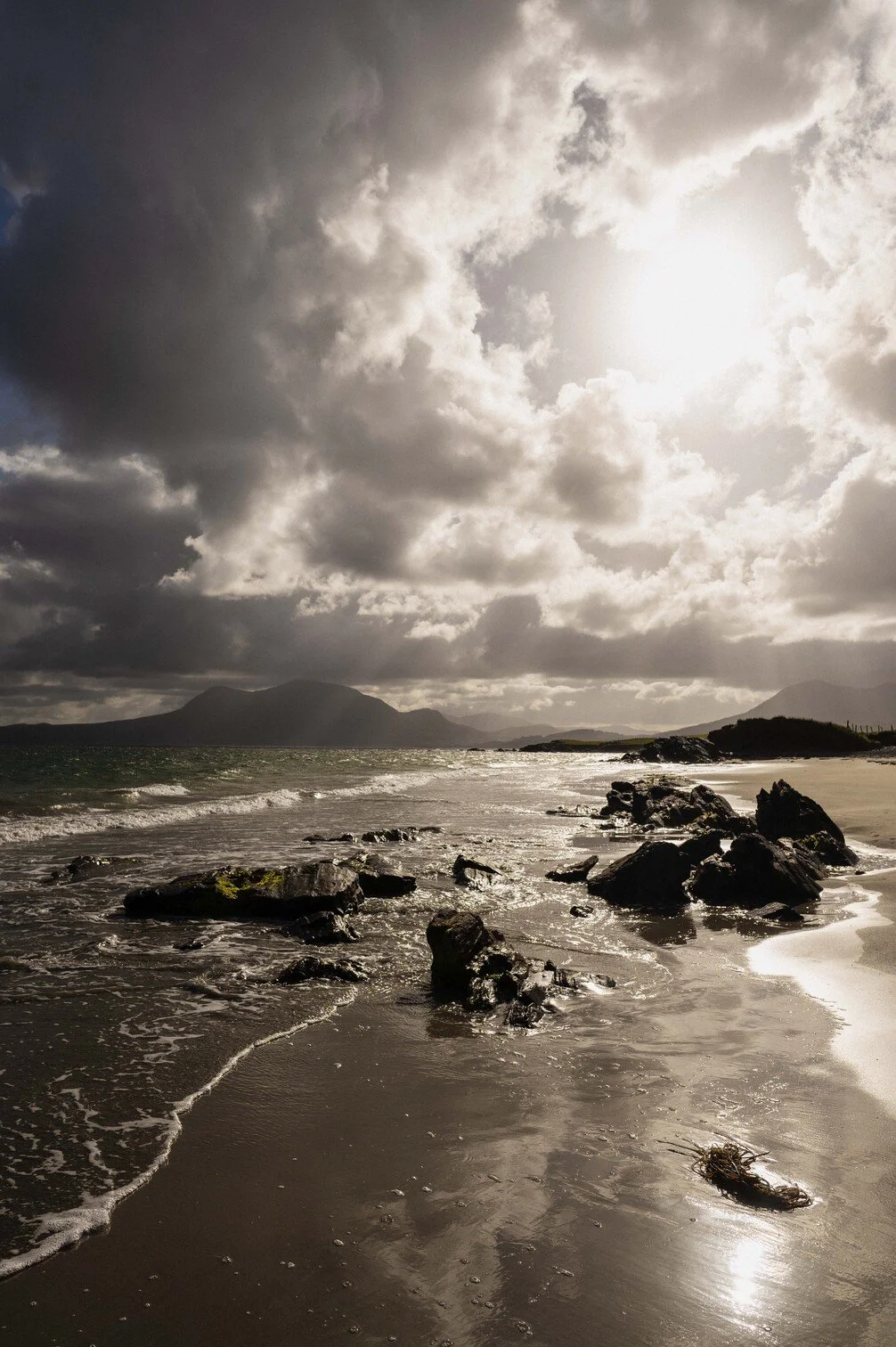 Een zee met rotsen op het strand onder een bewolkte hemel met de zon die schijnt gefotografeerd door Eric van den Elsen.