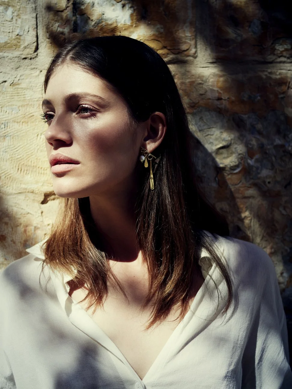 Close-up van een vrouw met bruin haar tegen een bakstenen muur, zonlicht op haar gezicht, wit blouse, oorbellen gefotografeerd door Eric van den Elsen.