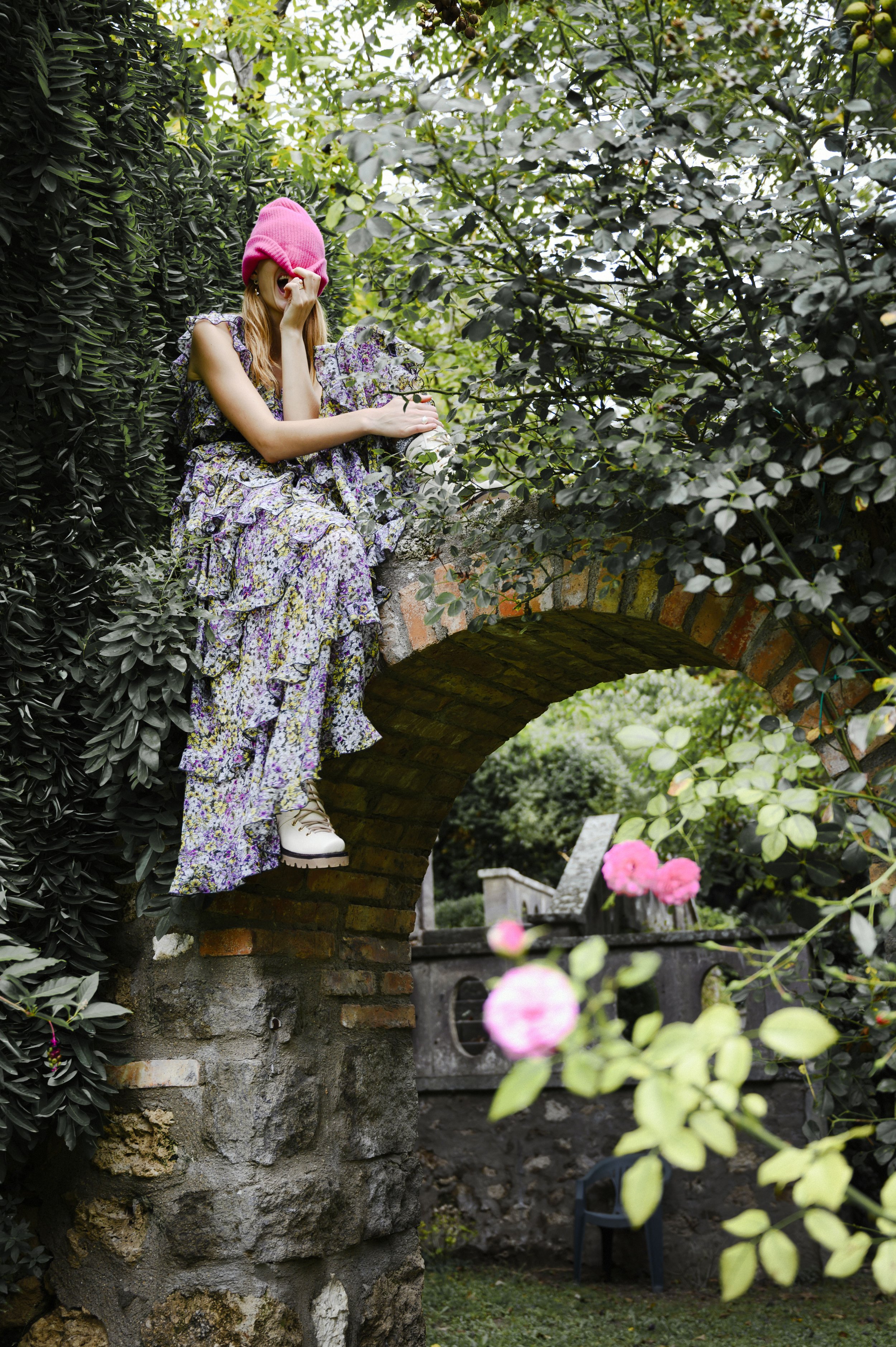 Een jonge vrouw zit op een stenen boog in een tuin, omgeven door groen en roze bloemen. Ze draagt een lange, bloemenjurk, witte sneakers, en een roze muts. Ze lacht en houdt haar hand bij haar gezicht gefotografeerd door Eric van den Elsen.