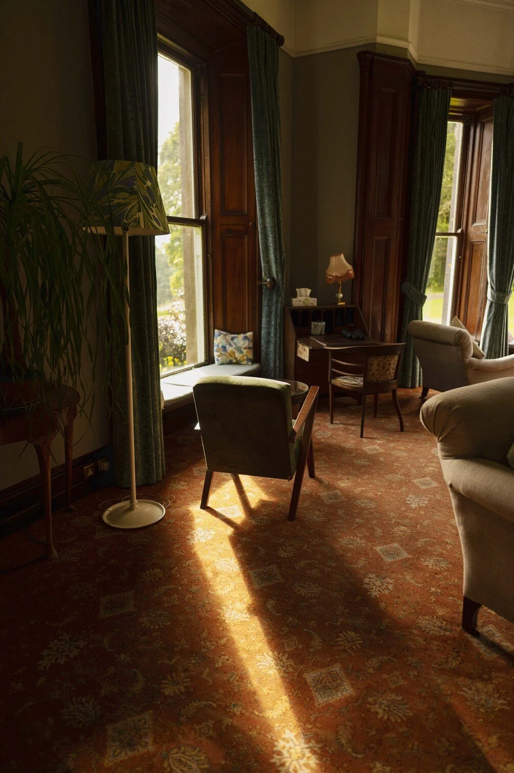 Interieur van een klassieke woonkamer met grote ramen, houten kozijnen, vintage meubels waaronder een groene fauteuil, een bureau en bank, met een vloerbedekking met oranje en gouden patronen gefotografeerd door Eric van den Elsen voor Vogue Living. 