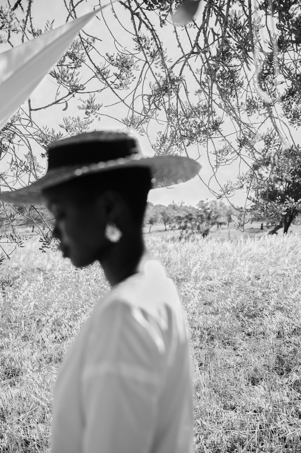 Een silhouet van een vrouw met een grote hoed, sieraden en een lichte jurk, staand in een veld met bomen en gras, onder een takkenwolk gefotografeerd door Eric van den Elsen.
