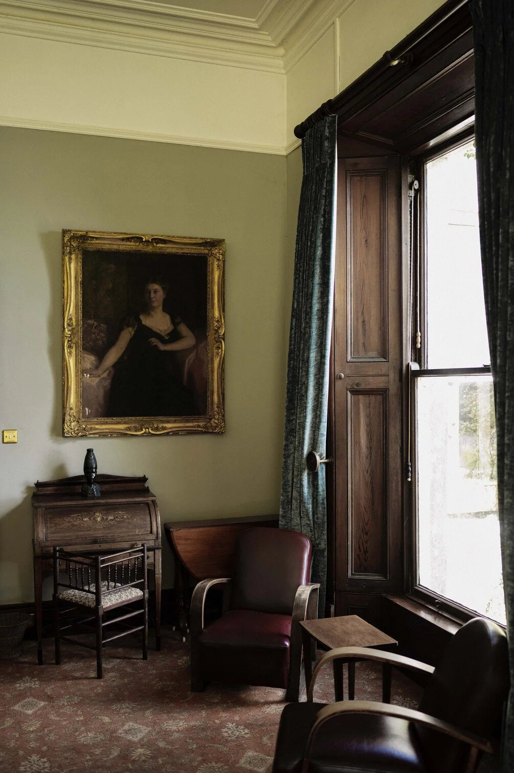 Een oude kamer met een grote raam, een moderne stoel en een klassiek bureau. Aan de muur hangt een groot portret van een vrouw in een zwarte jurk in een gouden lijst gefotografeerd door Eric van den Elsen.