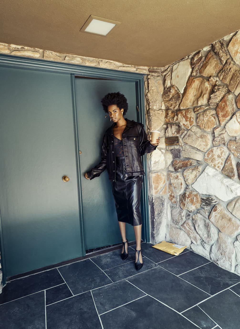 Een vrouw met kroeshaar staat voor een groene deur, gekleed in een zwart leren outfit en hoge hakken, en houdt een kopje koffie vast gefotografeerd door Eric van den Elsen.