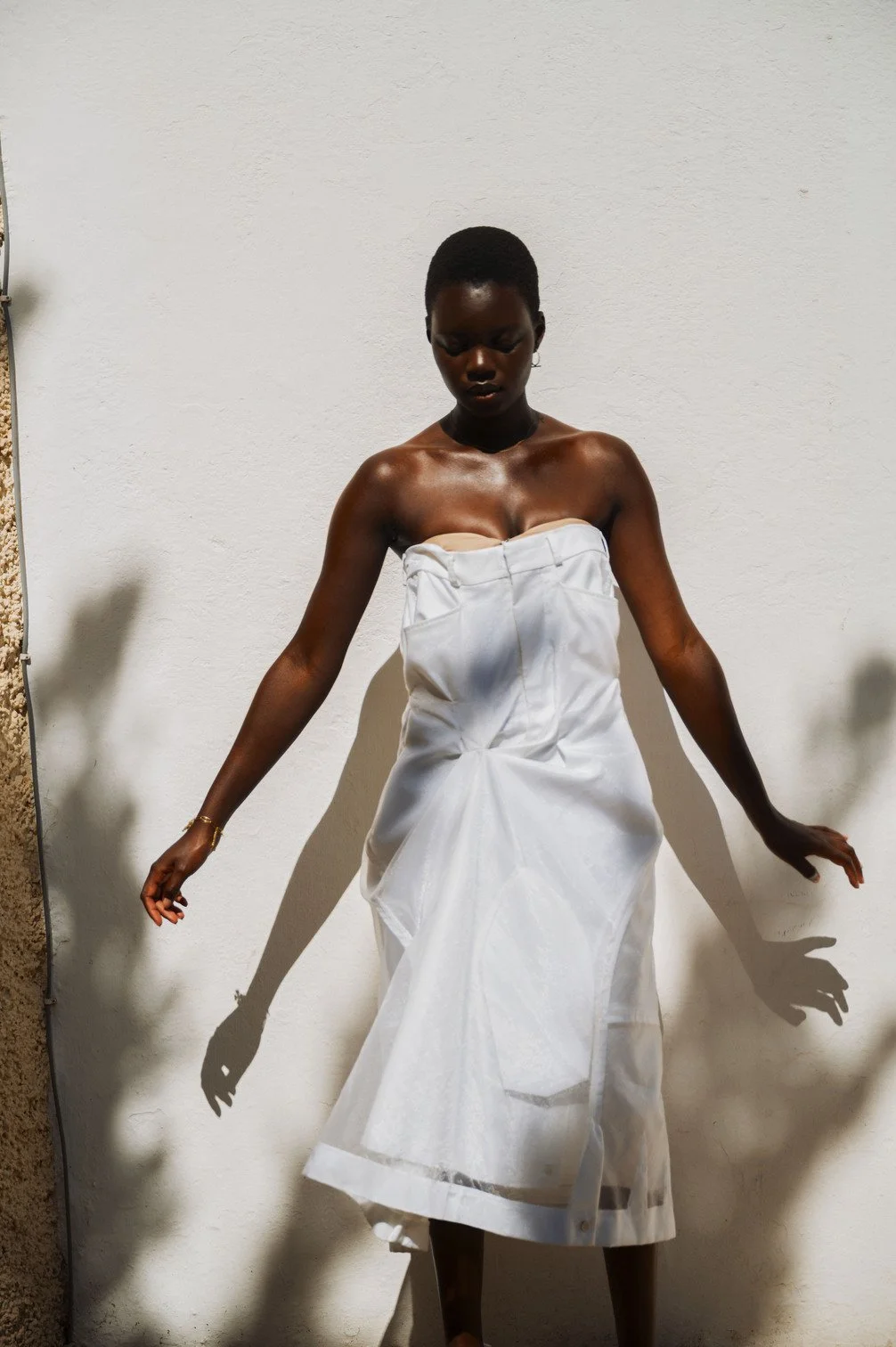 Een vrouw met een donkere huid en kort haar draagt een wit katoenen jurk en staat tegen een witte muur, waardoor haar schaduw zichtbaar is op de muur gefotografeerd door Eric van den Elsen.