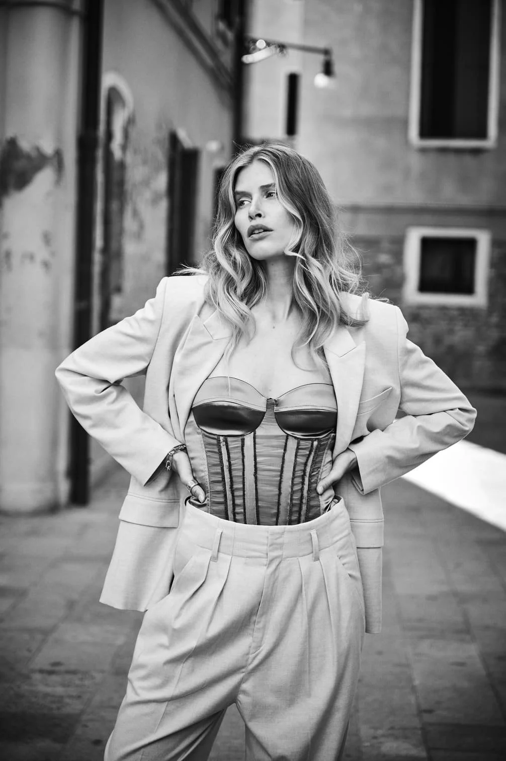 Zwarte en witte foto van een vrouw met golvend haar, gekleed in een blazer, een corset en brede broek, staand op straat met een gebouw op de achtergrond gefotografeerd door Eric van den Elsen.