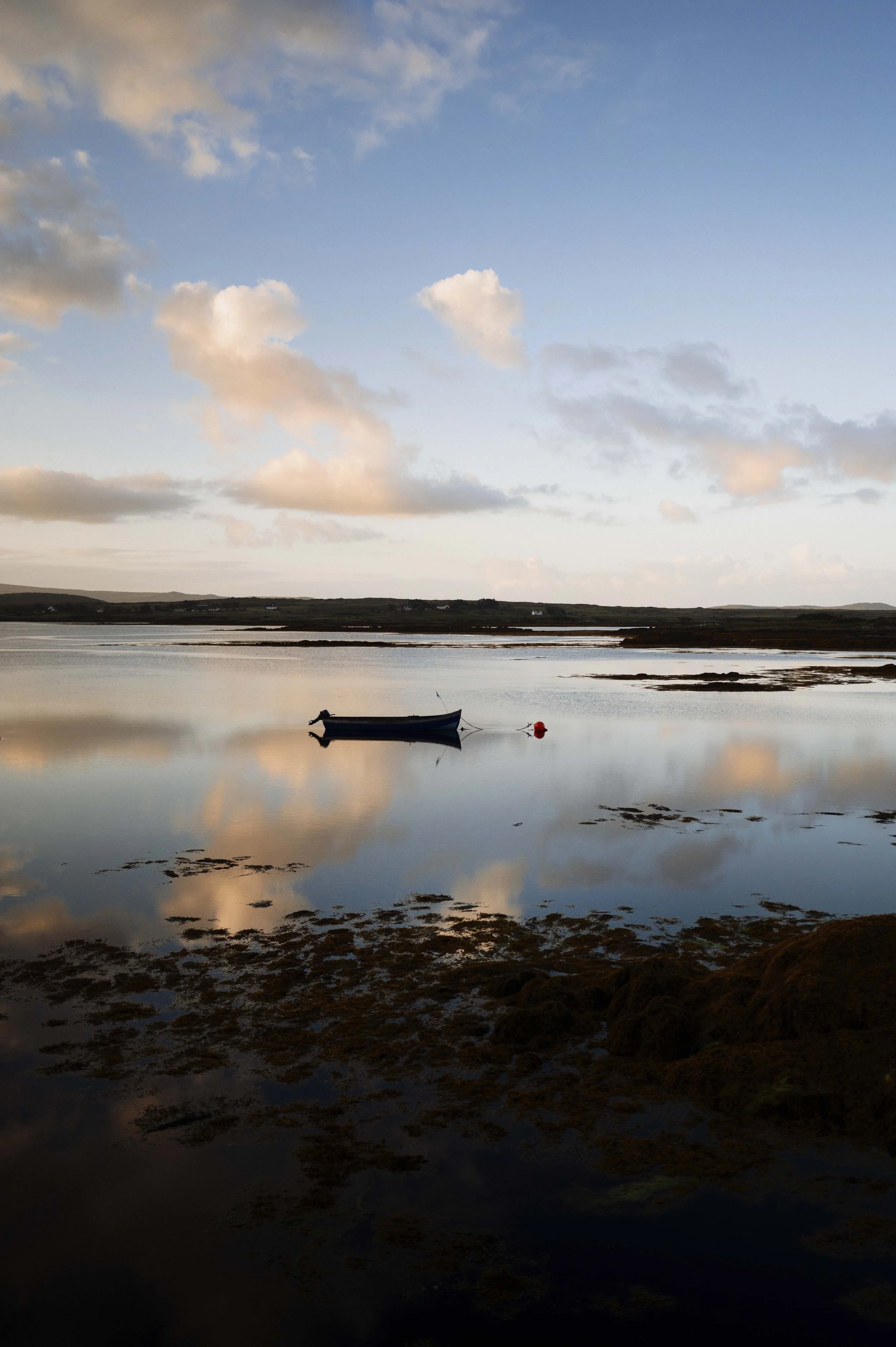 Een rustige zee met een klein bootje dat vast ligt aan een rode boei, onder een bewolkte hemel met reflectie in het water gefotografeerd door Eric van den Elsen voor Vogue Living.