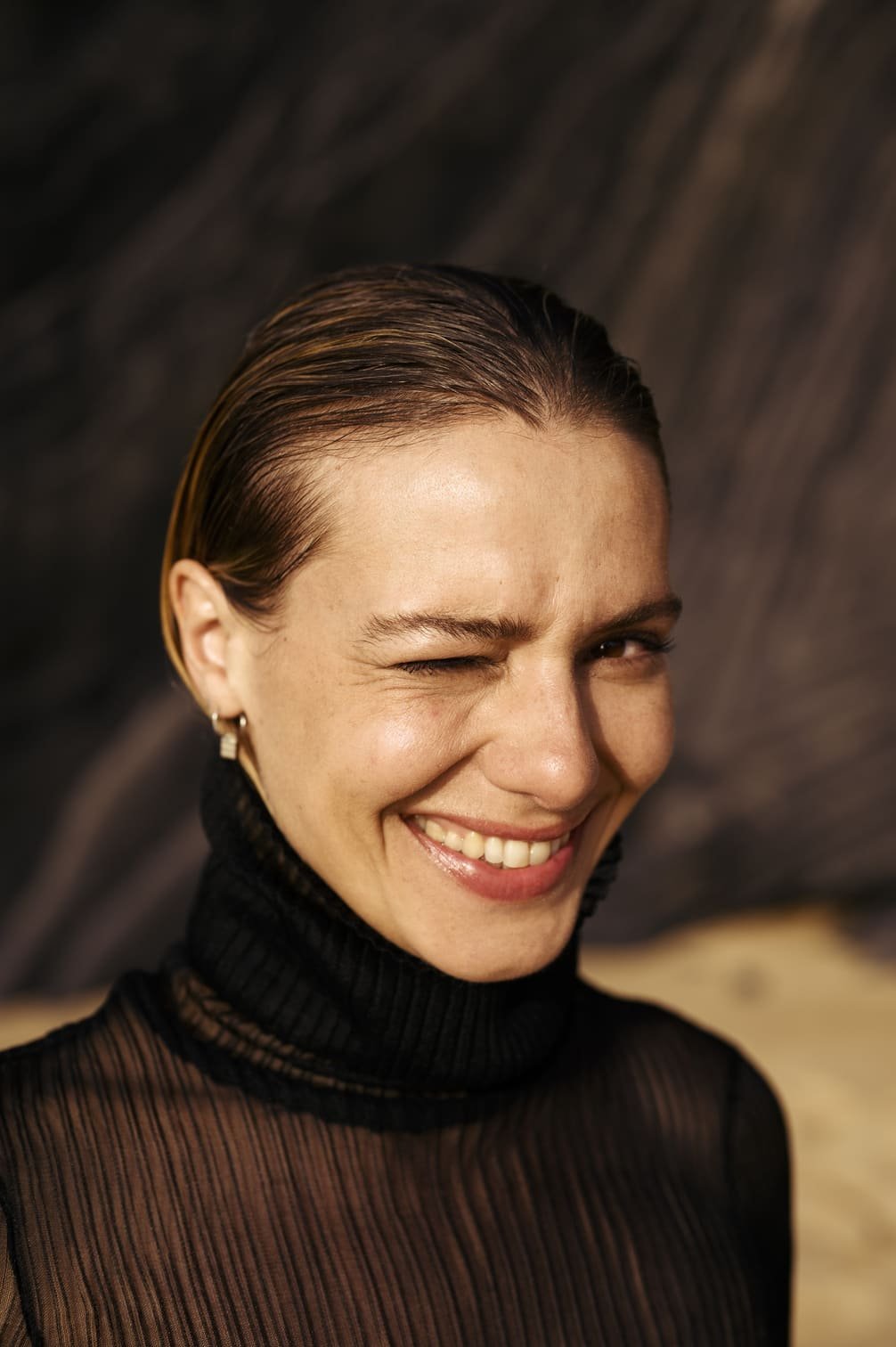 Sallie Harmsen lachend en knippert met één oog, met nat haar en een zwarte getailleerde top gefotografeerd door Eric van den Elsen.