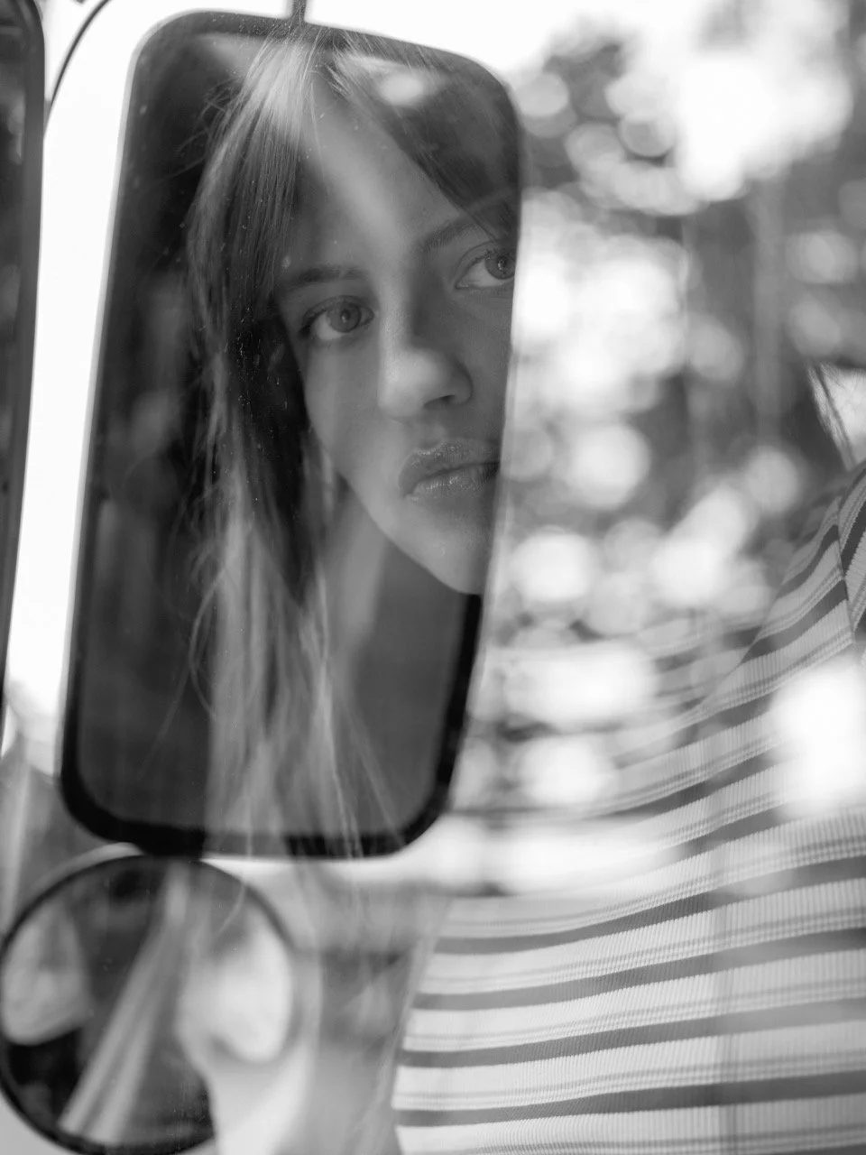 Close-up van een vrouw die in een auto naar haar spiegel kijkt, zwart-wit foto, gedeeltelijke spiegelreflectie, met een achtergrond van boombladeren gefotografeerd door Eric van den Elsen.