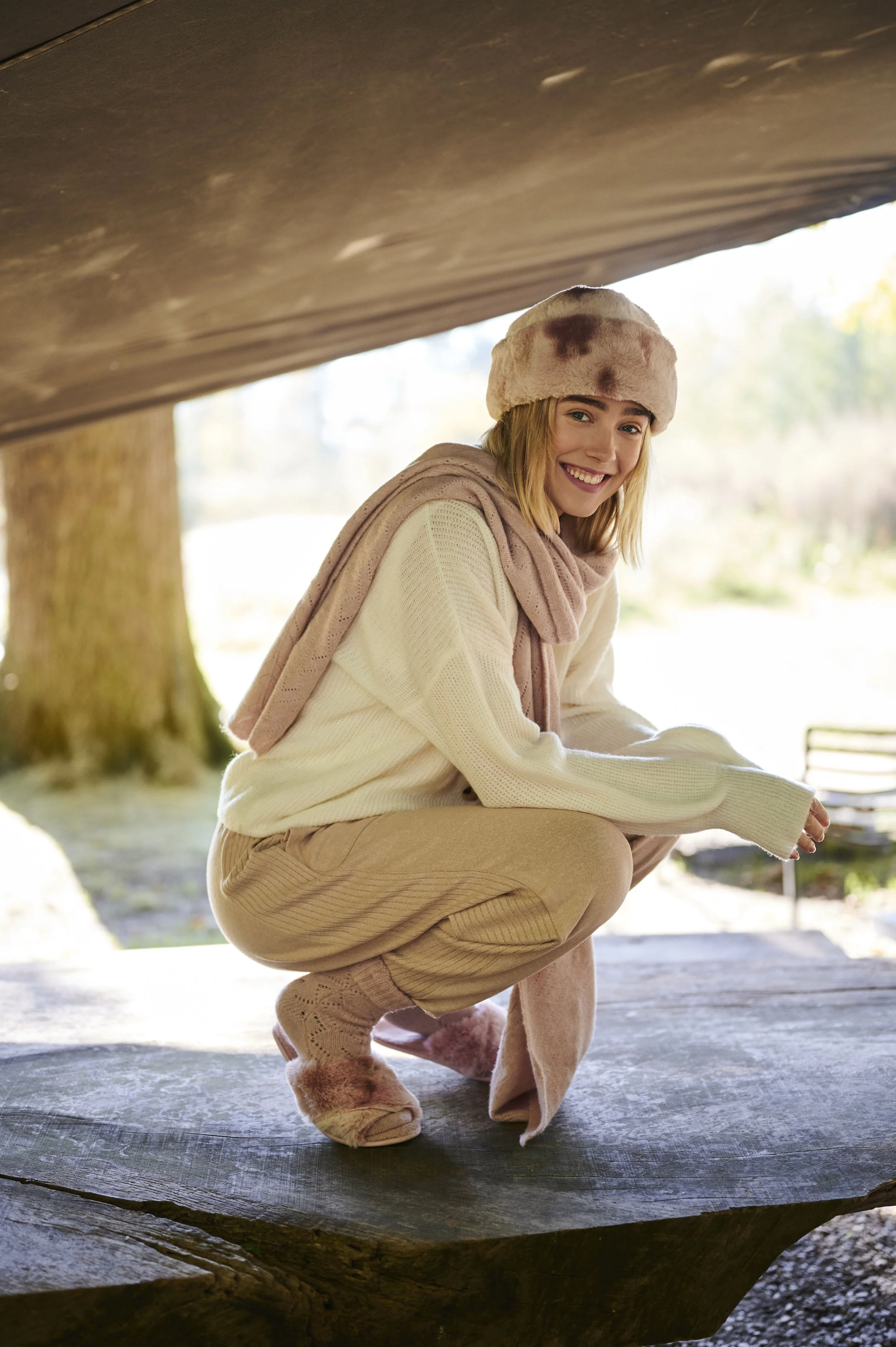 Vrouw in winterkleding die knielt op een boomstam onder een beschaduwde bogen, lachend naar de camera gefotografeerd door Eric van den Elsen voor Bart Amsterdam.