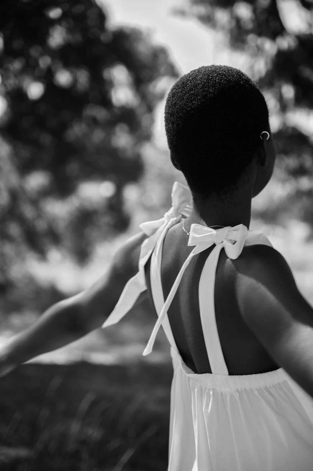 Een persoon met kort haar en een zijaanzicht, gekleed in een witte jurk met strikdetails op de schouders, staat buiten met een blurry groene achtergrond gefotografeerd door Eric van den Elsen.