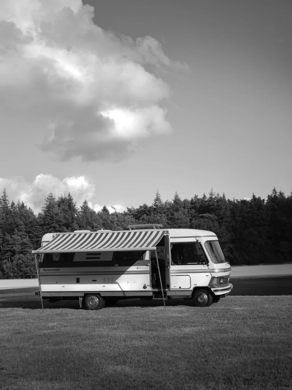 Een camperpark op een grasveld met bomen op de achtergrond en een bewolkte hemel erboven, in zwart-wit gefotografeerd door Eric van den Elsen.