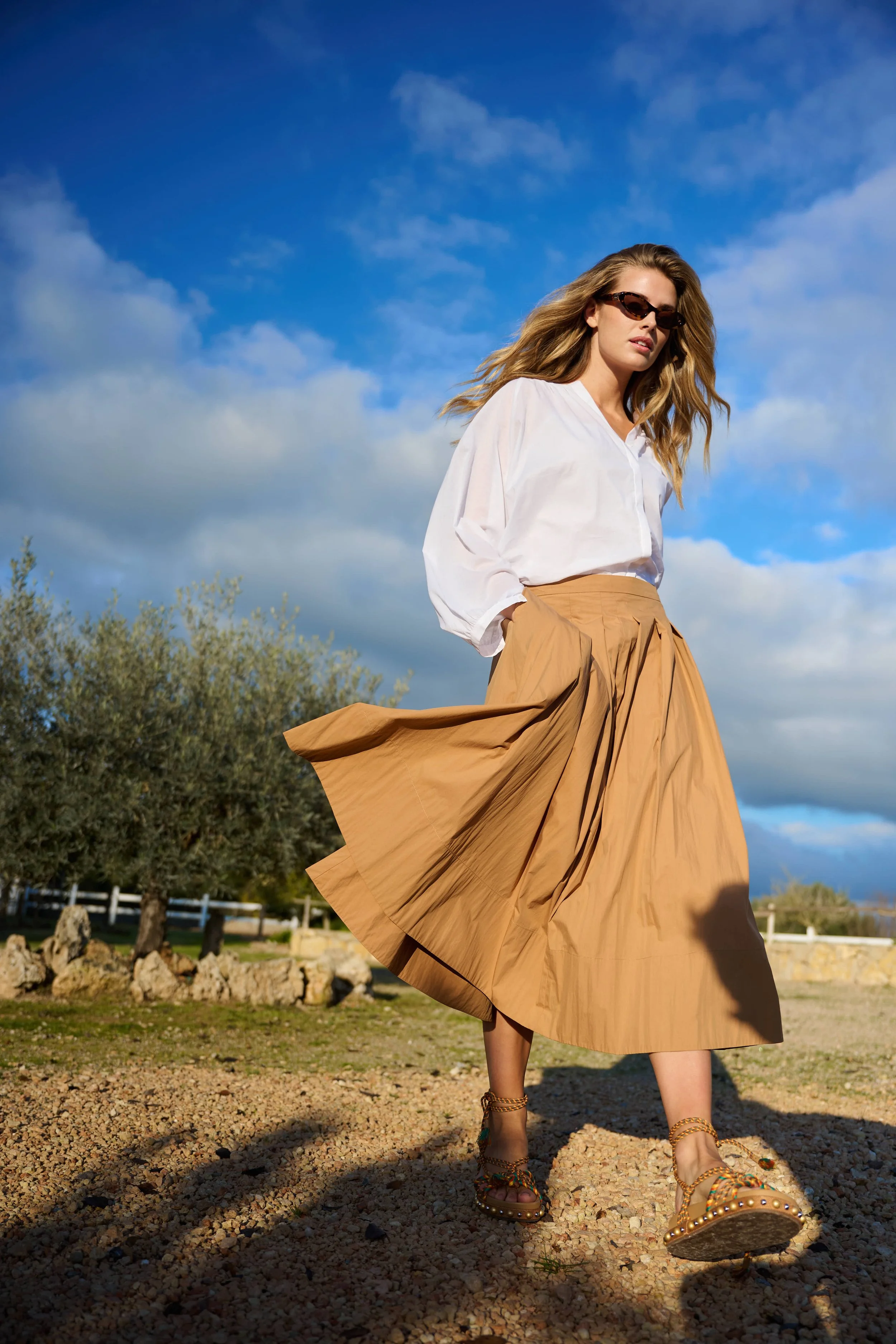 Een vrouw in een witte blouse en een lange beige rok wandelen op een gravelpad onder een blauwe lucht met wolken, draagt sandalen en zonnebril gefotografeerd door Eric van den Elsen voor Josephine & Co.