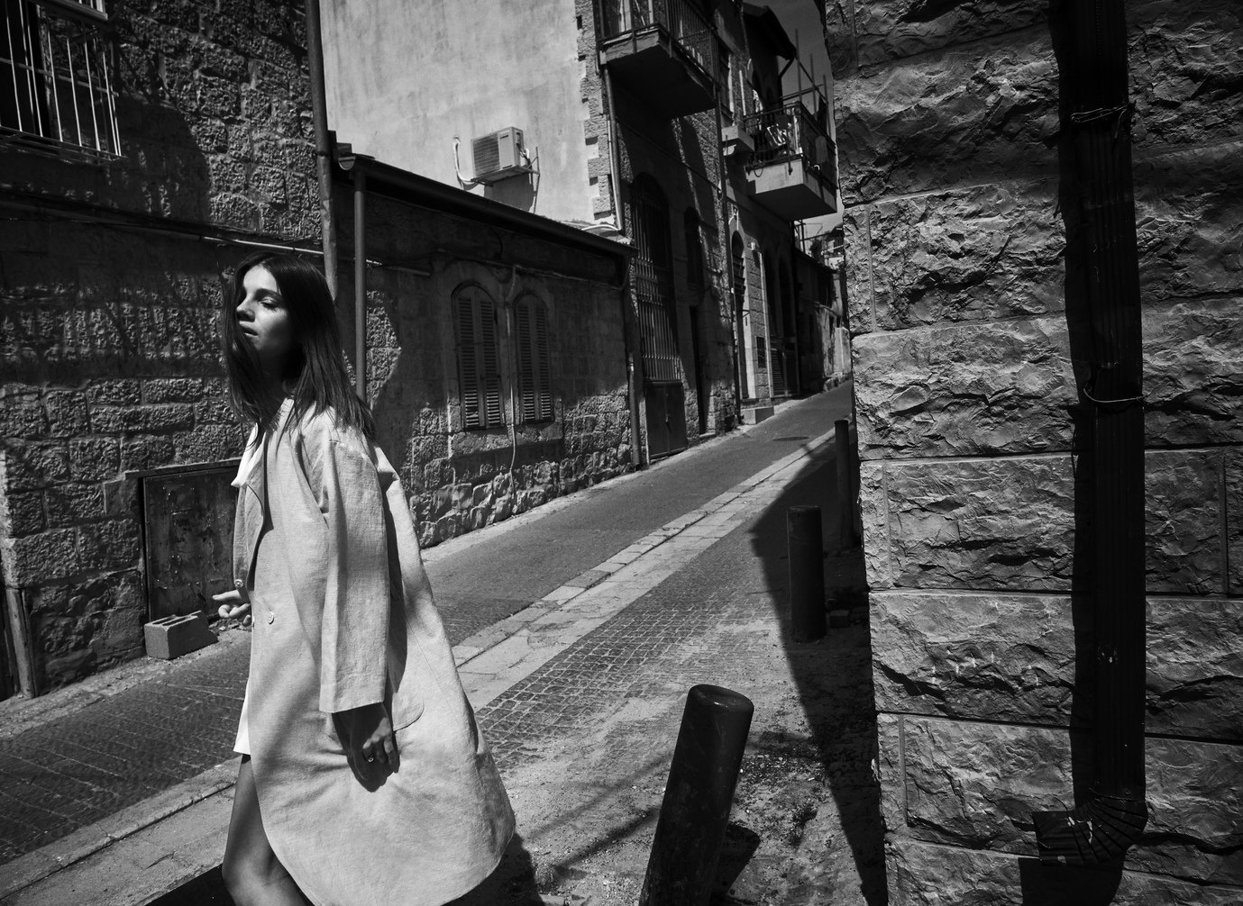 Een vrouw met donker haar en een lange jas staat op een straat in een stadswijk, omringd door oude stenen gebouwen en schaduwrijke omgeving gefotografeerd door Eric van den Elsen.