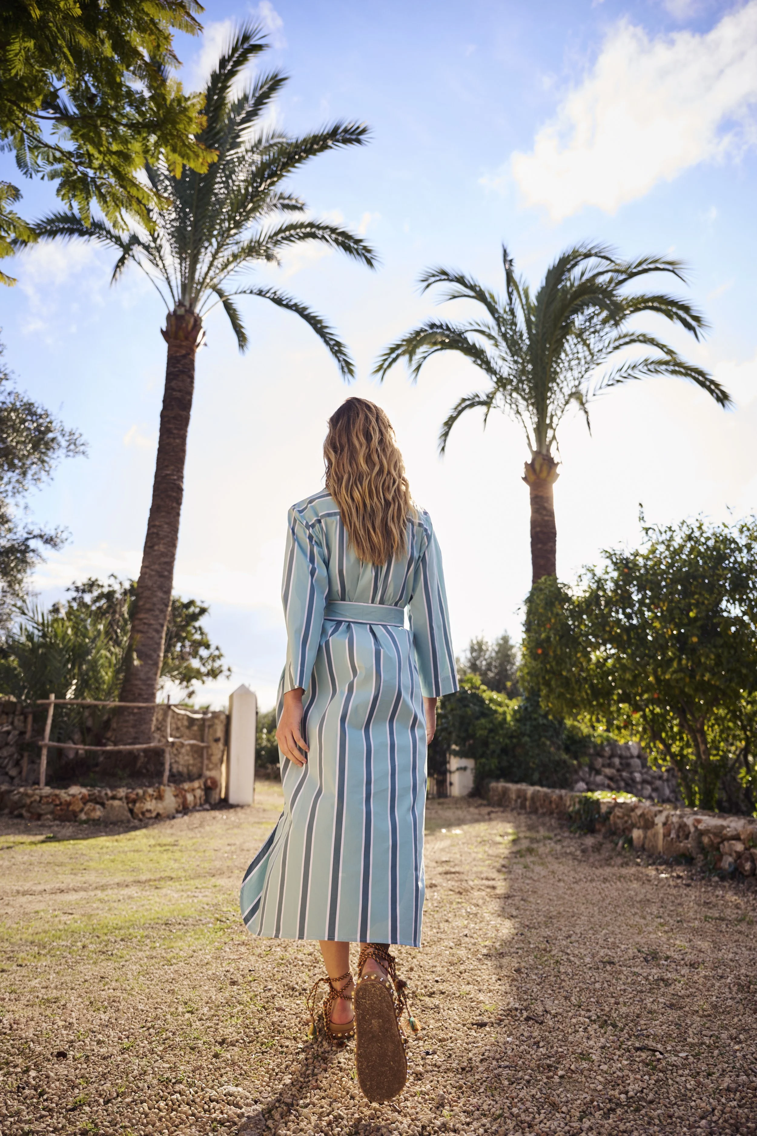 Vrouw in een lange, gestreepte jurk loopt weg op een zandweg onder palmbomen, met een heldere blauwe lucht en enkele wolken gefotografeerd door Eric van den Elsen voor Josephine & Co.