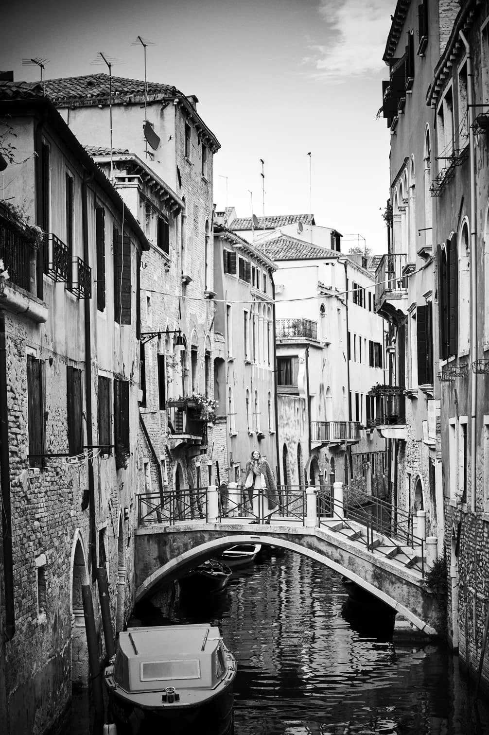 Zwart-wit foto van een smal kanaal omringd door oude gebouwen met kleine balkons en ramen, een brug over het kanaal met een persoon die eroverheen loopt, en een klein bootje in het water gefotografeerd door Eric van den Elsen.