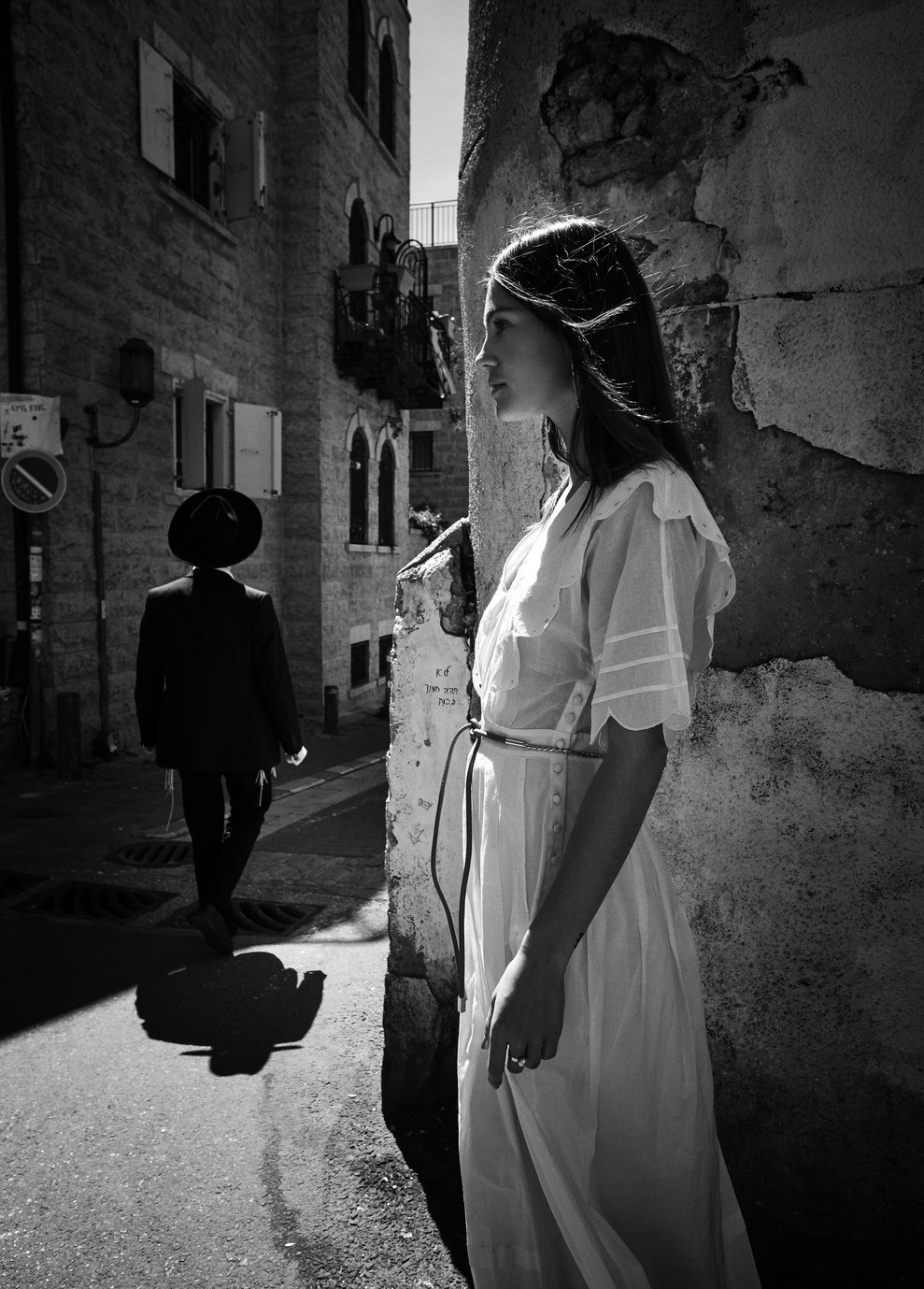Een zwart-wit foto van een jonge vrouw in een witte jurk die tegen een oude stenen muur leunt, met een man in een pak en hoed die wegloopt op een smalle straat gefotografeerd door Eric van den Elsen.