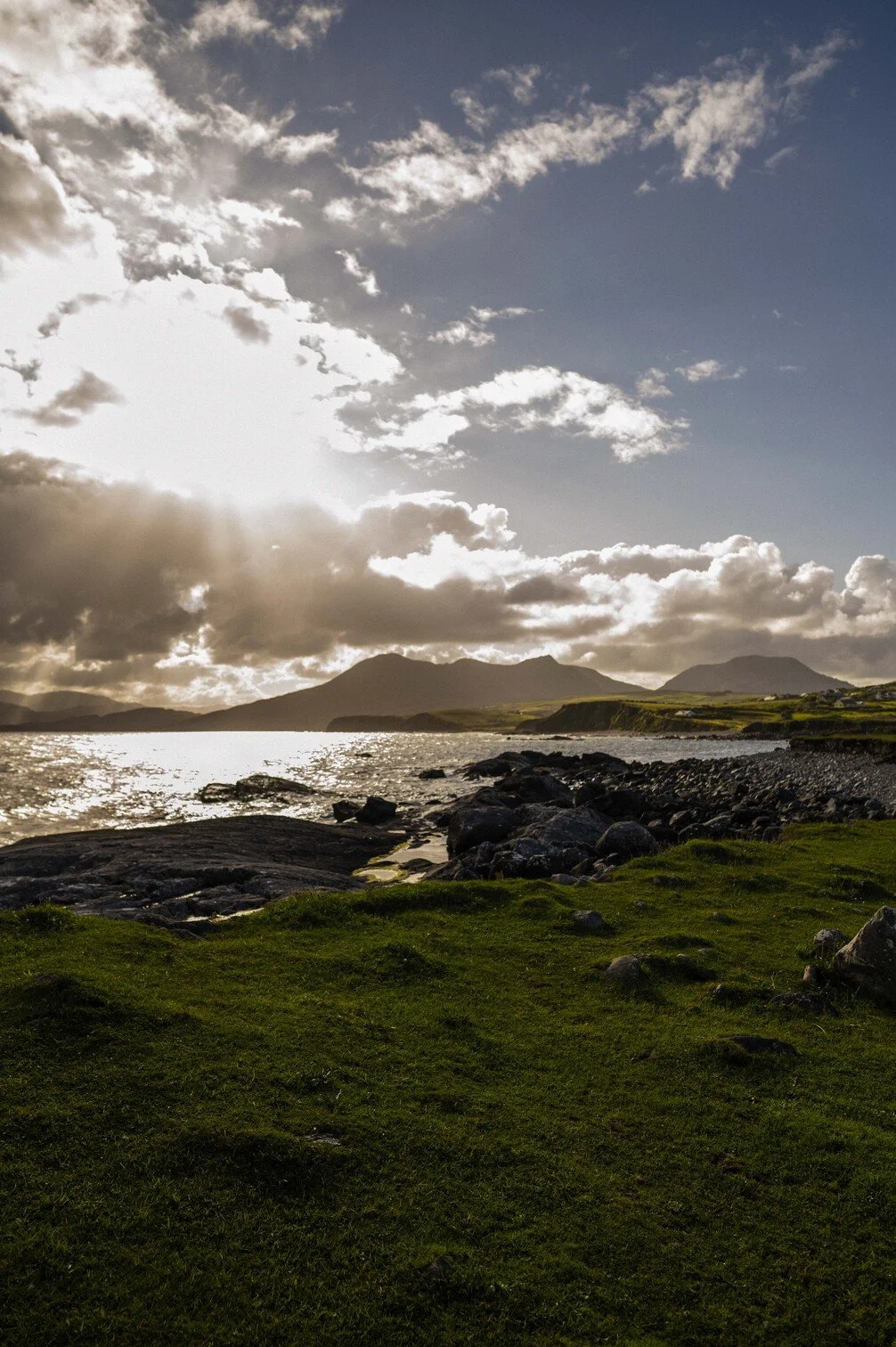 Een kustlandschap met groene grasvelden, rotsen langs de waterkant, een oceaan, en bergen op de achtergrond onder een bewolkte hemel met de zon die door de wolken schijnt gefotografeerd door Eric van den Elsen.
