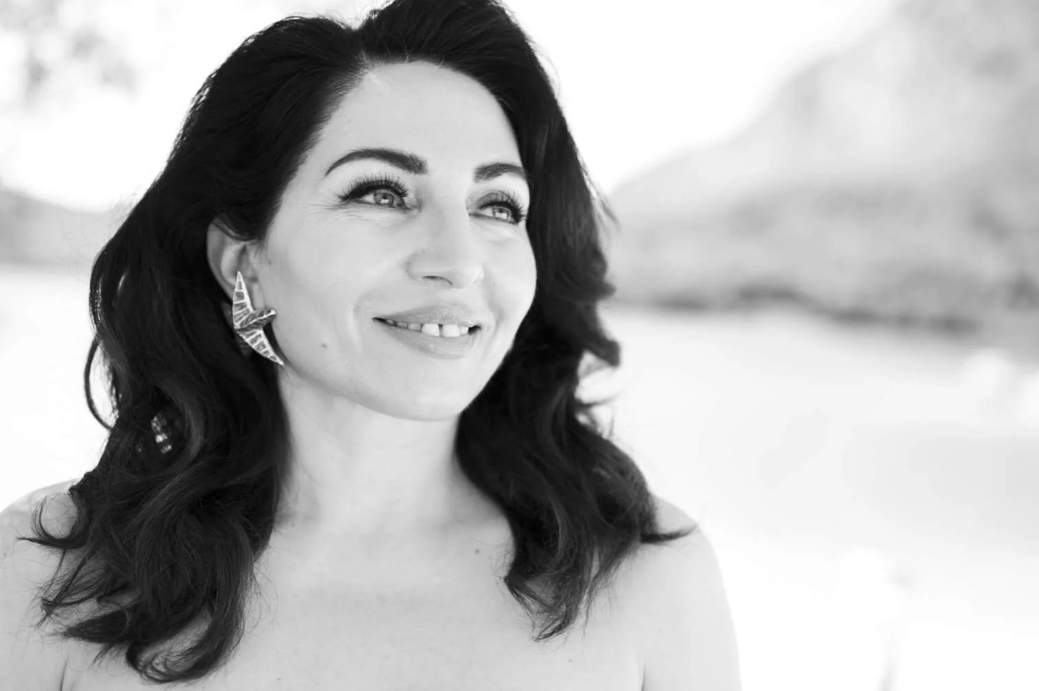 Zwart-wit foto van een glimlachende Nazmiye Oral met lang, golvend haar en grote oorbellen, buiten met een berg op de achtergrond gefotografeerd door Eric van den Elsen.