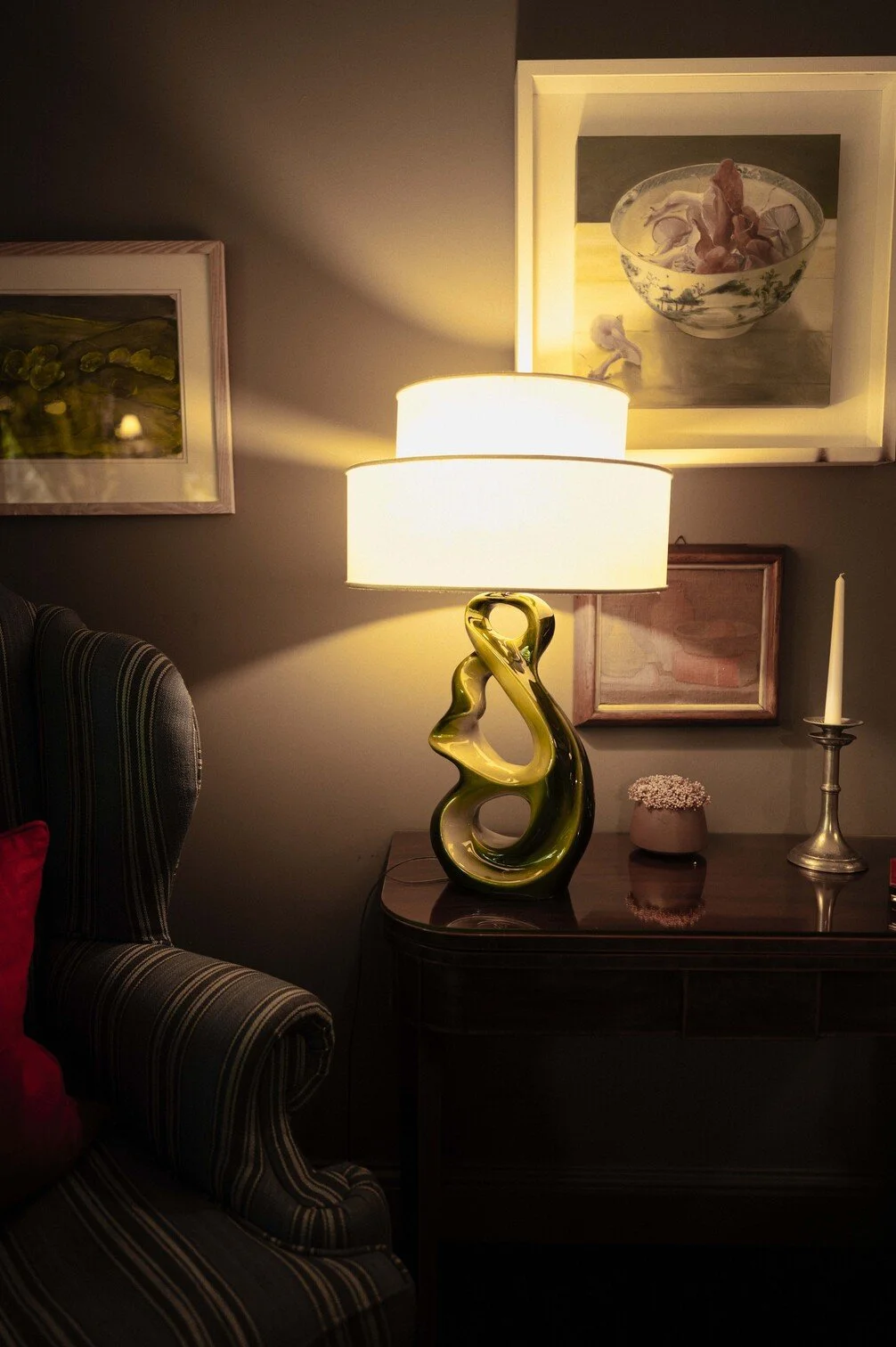 Een landelijke woonkamer met een gestreepte fauteuil, een tafel met een moderne groene glaslamp, een vaas met bloemen en een kandelaarkandel op een donkerhouten kast, en schilderijen aan de muur gefotografeerd door Eric van den Elsen.