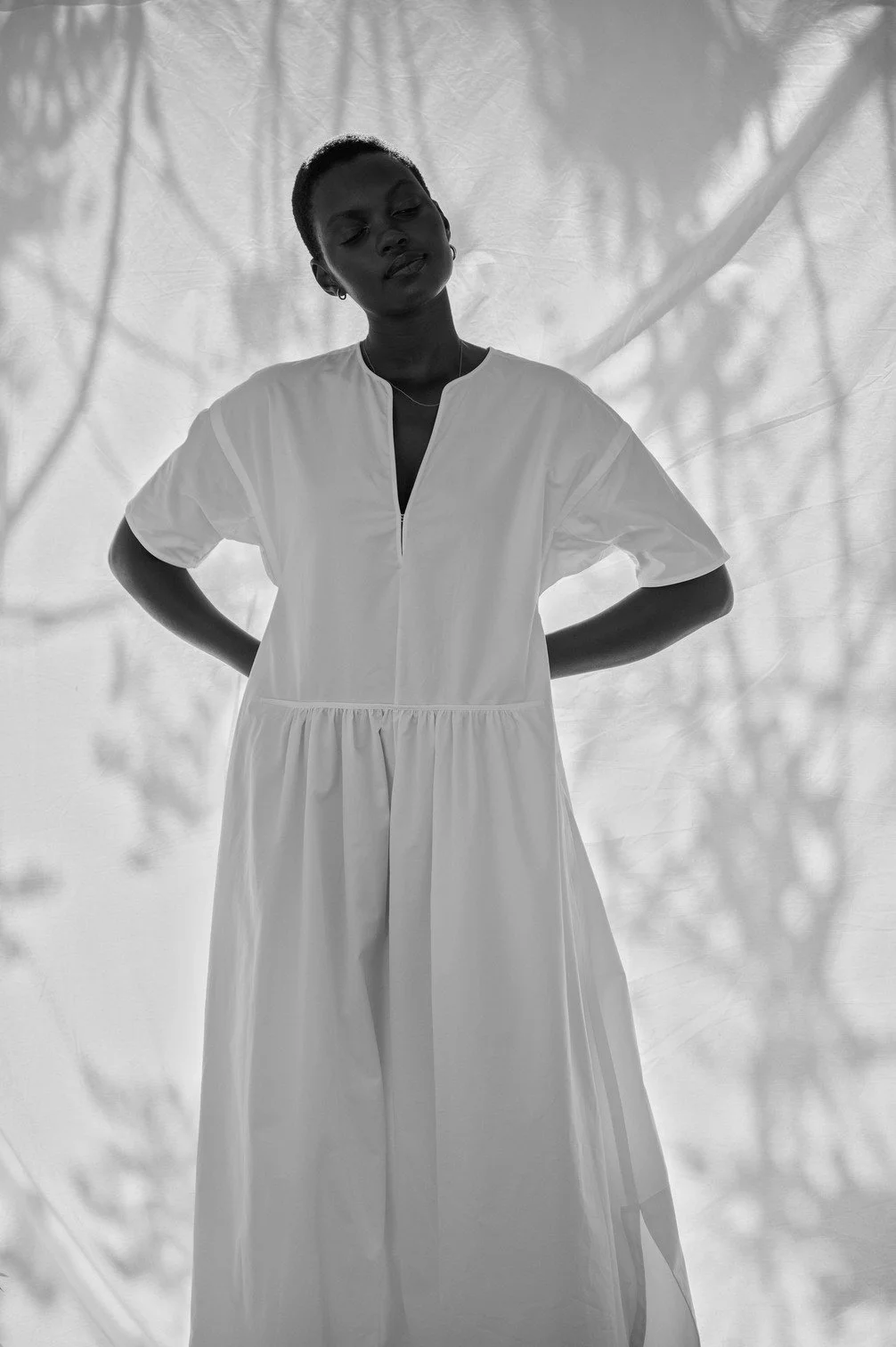 Een vrouw in een witte jurk staat met haar handen in haar heupen voor een lichte achtergrond met schaduwen gefotografeerd door Eric van den Elsen.