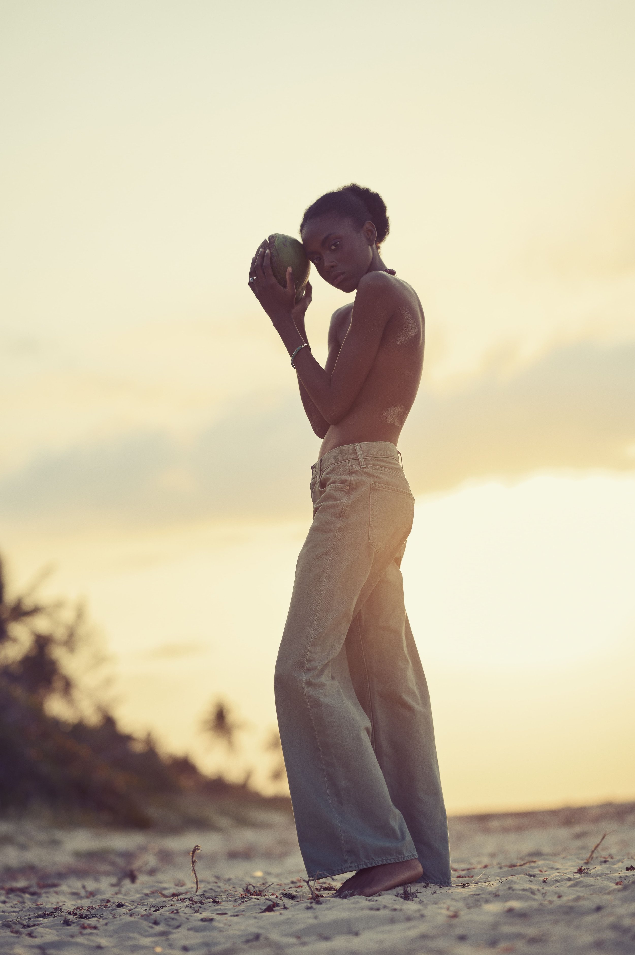 Een jonge man zonder shirt, die op een zandstrand staat, een kokosnoot vasthoudt en naar de camera kijkt bij zonsondergang gefotografeerd door Eric van den Elsen.