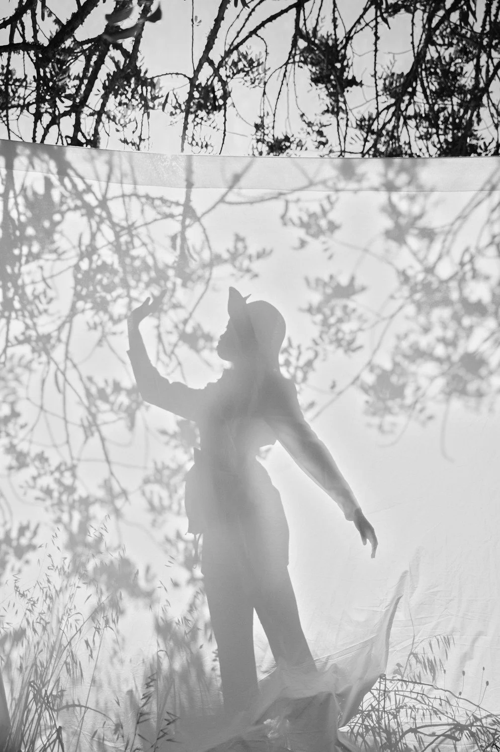 Silhouet van een vrouw die met haar arm omhoog staat, het lijkt op een filmset of artistieke decor in de natuur met bomen en planten op de achtergrond gefotografeerd door Eric van den Elsen.