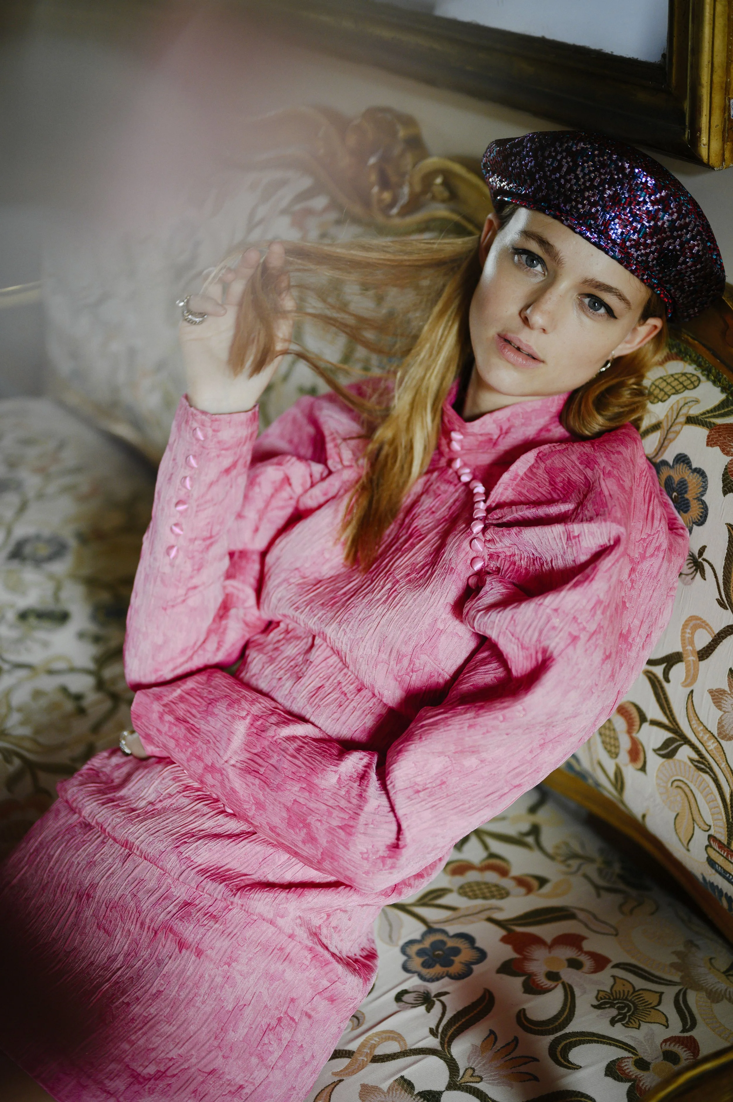 Een jonge vrouw met rood haar, gekleed in een roze jurk met puffermouwen, ligt op een bank met een vogelprint en kijkt naar de camera gefotografeerd door Eric van den Elsen.