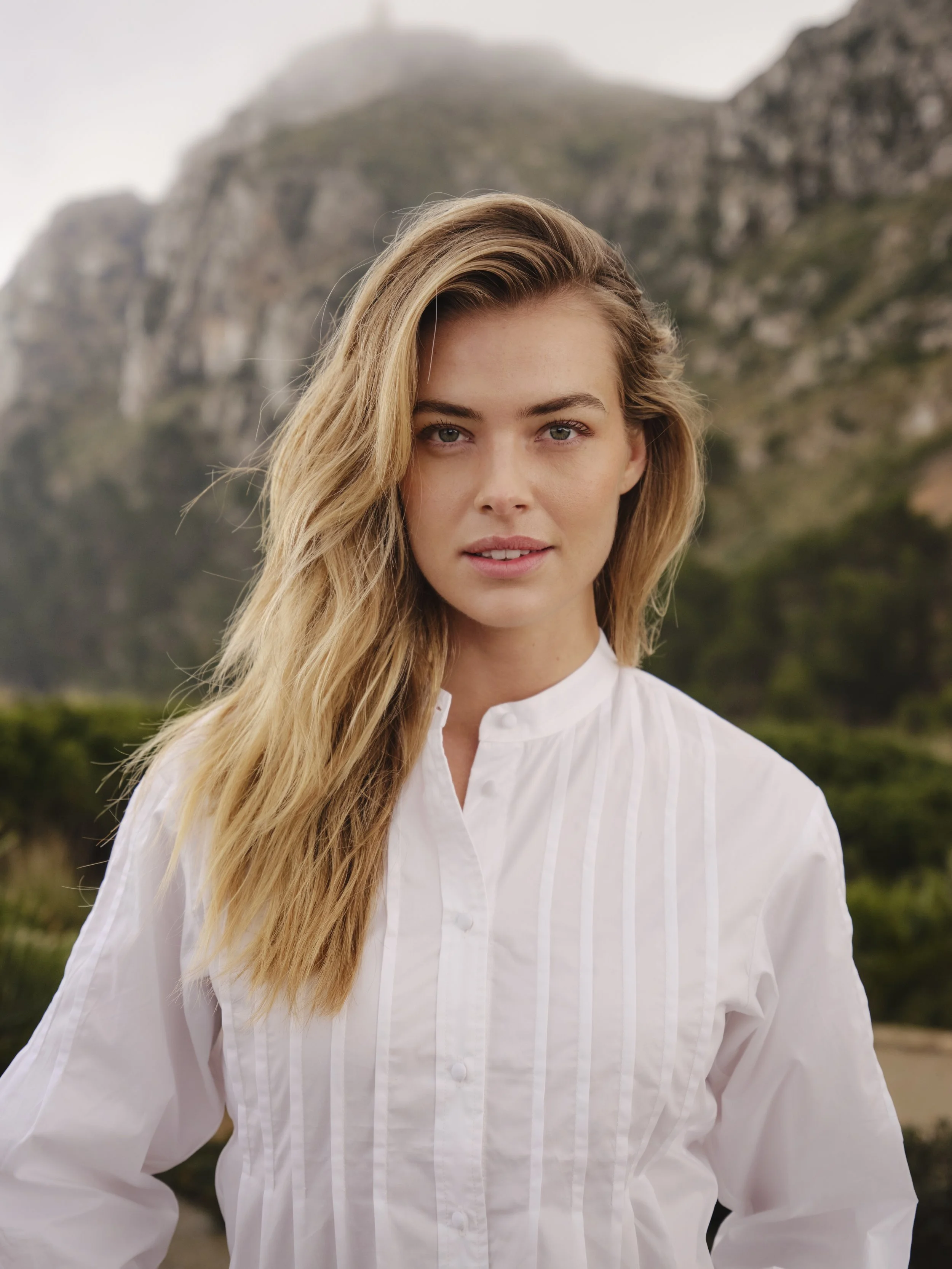 Portret van een jonge vrouw met lang blond haar, witte blouse en een natuurlijke achtergrond van bergen en groen landschappen gefotografeerd door Eric van den Elsen voor Josephine & Co.